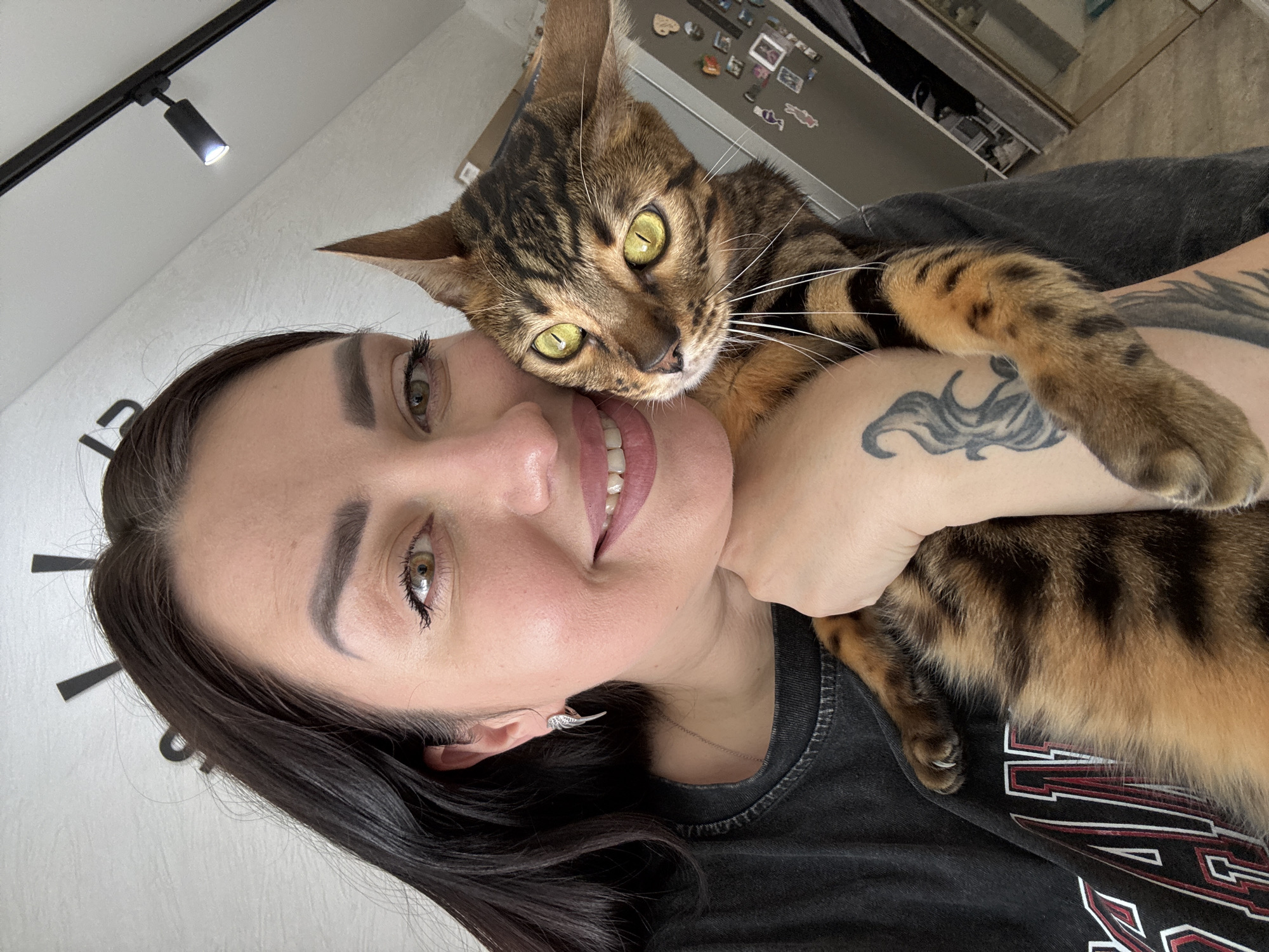 A selfie-style portrait features a woman and a Bengal cat in an indoor setting. The woman, with dark hair, dark eyebrows, and light brown/hazel eyes, smiles directly at the camera, revealing her teeth. She wears a dark t-shirt with a partially visible graphic design featuring white and red elements. A tattoo resembling stylized waves or tentacles is visible on her left upper arm. A small, silver-colored earring shaped like a wing is in her left ear.
Perched on her shoulder and chest is a Bengal cat, characterized by its distinctive brown and black striped and rosetted fur pattern, and striking bright green-yellow eyes. The cat's head is close to the woman's face, and it looks directly into the camera. One of its paws rests gently on the woman's chest.
The background suggests a home environment. To the upper left, a white ceiling features a black track light fixture with a lit spotlight. On the white wall behind the woman, black minimalist markers, likely part of a large wall clock, are visible. In the upper right background, a metallic surface, possibly a refrigerator or cabinet, is partially visible, adorned with various small magnets or photos. The overall impression is one of affectionate companionship. No clear, legible text is visible in the image.