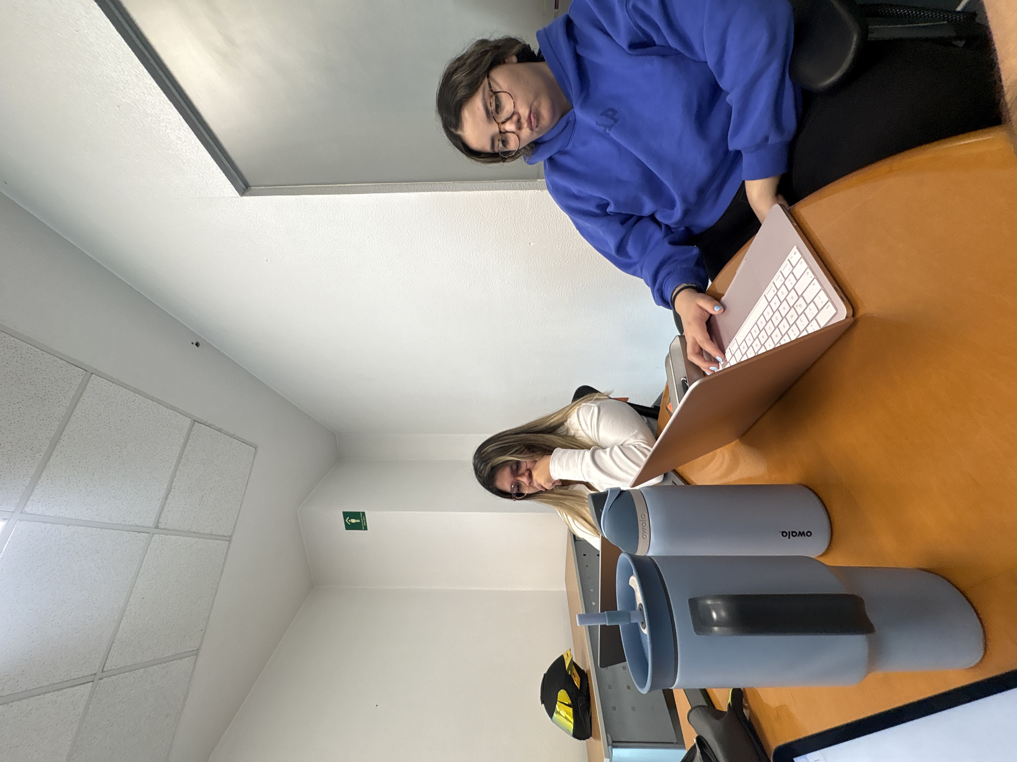 The photo depicts an interior scene, likely in a study area or library in Puebla, Mexico. Two young women are seated at a wooden table, each with a laptop. The woman on the right wears a blue hoodie and glasses, actively typing on her laptop. The other woman has long, blonde hair and is wearing a white long-sleeved shirt, appearing to be resting her face in her hands. Two light blue Owala water bottles are on the table. A black motorcycle helmet and other personal items are also present. The room has white walls and a drop ceiling. An emergency exit sign is visible on the wall.