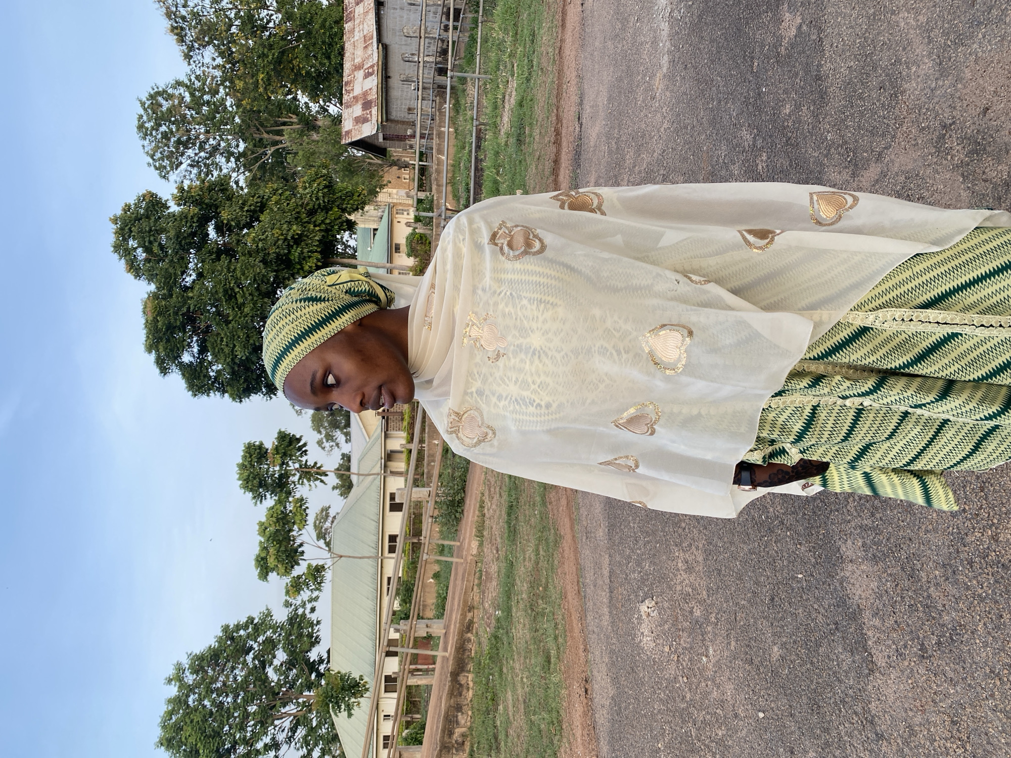 A woman stands on an asphalt road. She is wearing a green and yellow patterned dress and head covering, draped with a cream-colored shawl patterned with gold heart designs. The woman is looking down. Behind her, there is a building with a green roof, a fence, and some trees. In the background, there is also a metal shed-like structure. This is a shot taken at a site in Vom, Nigeria.