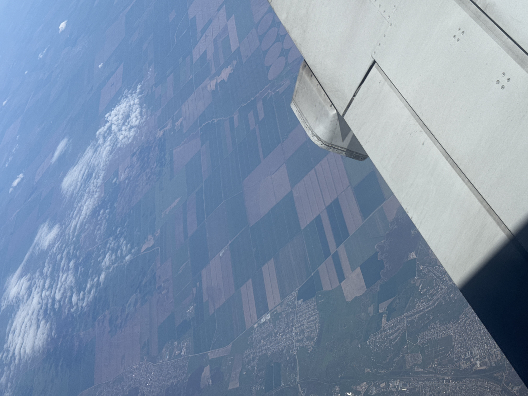 The image depicts an aerial view from an airplane window. The right side of the frame is dominated by a portion of the airplane's wing, colored light gray. Below the wing, the landscape is divided into a patchwork of agricultural fields, varying in color and texture, suggesting different crops or stages of growth. Further in the distance, towards the bottom of the frame, is a city, Забегалово, Russia. It appears densely populated, with a network of roads and buildings visible. Above the landscape is a layer of clouds.