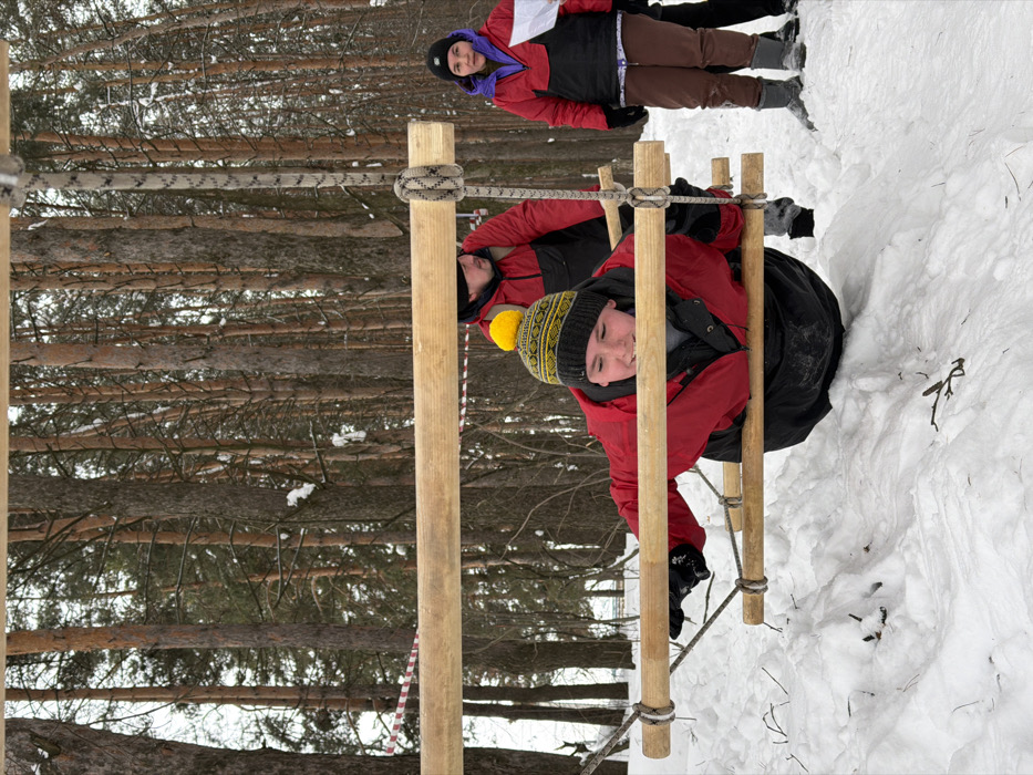 The image depicts a winter outdoor scene in a snowy forest in Staryi Senteg, Russia. A young individual, wearing a red and black jacket, black pants, black gloves, and a yellow-pompommed patterned knit hat, is actively navigating a low-lying obstacle. This obstacle consists of several horizontal wooden rungs suspended by ropes, with the individual positioned horizontally, passing through the rungs with their chest and abdomen supported. The ground is covered in deep, white snow with visible footprints and scattered twigs. In the mid-ground, to the right, an adult female stands wearing a red and black jacket, brown pants, light-colored boots, and a black beanie, holding a white piece of paper. Behind the central figure, a third individual, an adult male in a red and black jacket and a dark hat, is partially visible. The background is dominated by a dense growth of tall, slender trees, indicative of a pine forest, with some snow clinging to branches. A red and white striped tape, likely a course marker, is visible horizontally in the upper mid-ground. The lighting is overcast, creating diffuse shadows.