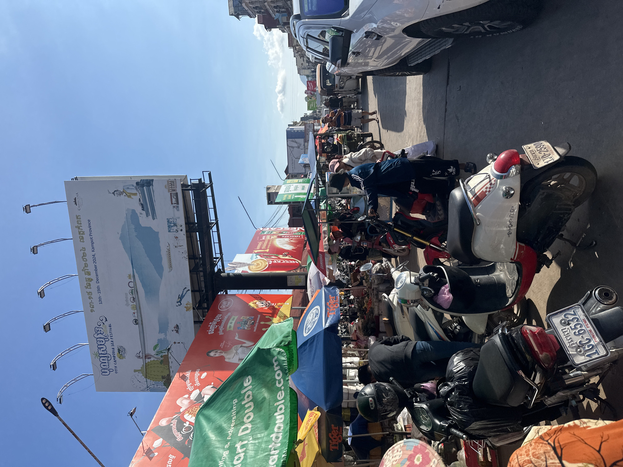 This is a daytime, eye-level, outdoor, street-level photograph taken in Kampot, Cambodia. The scene depicts a busy street market with numerous stalls and vehicles. In the foreground, several motorcycles are parked, some with people standing near them. One motorcycle has a license plate that reads "1GC-6532 PHNOM PENH". A white pickup truck is parked on the right side of the street, partially obscuring the view of the road.
In the midground, market stalls with colorful umbrellas are set up, selling various goods. People are seen browsing and interacting at the stalls. A person in a blue jacket and black pants appears to be walking through the market. In the background, there are buildings and more market stalls extending down the street.
Dominating the upper portion of the image is a large billboard advertising the "11th Cambodia Sea Festival" taking place from "13th - 15th December 2024, Kampot Province". The billboard features illustrations and text in both Khmer and English. Other smaller billboards and signs are also visible, advertising products and services. The sky is clear and blue with some scattered clouds. The overall atmosphere suggests a vibrant and active marketplace.