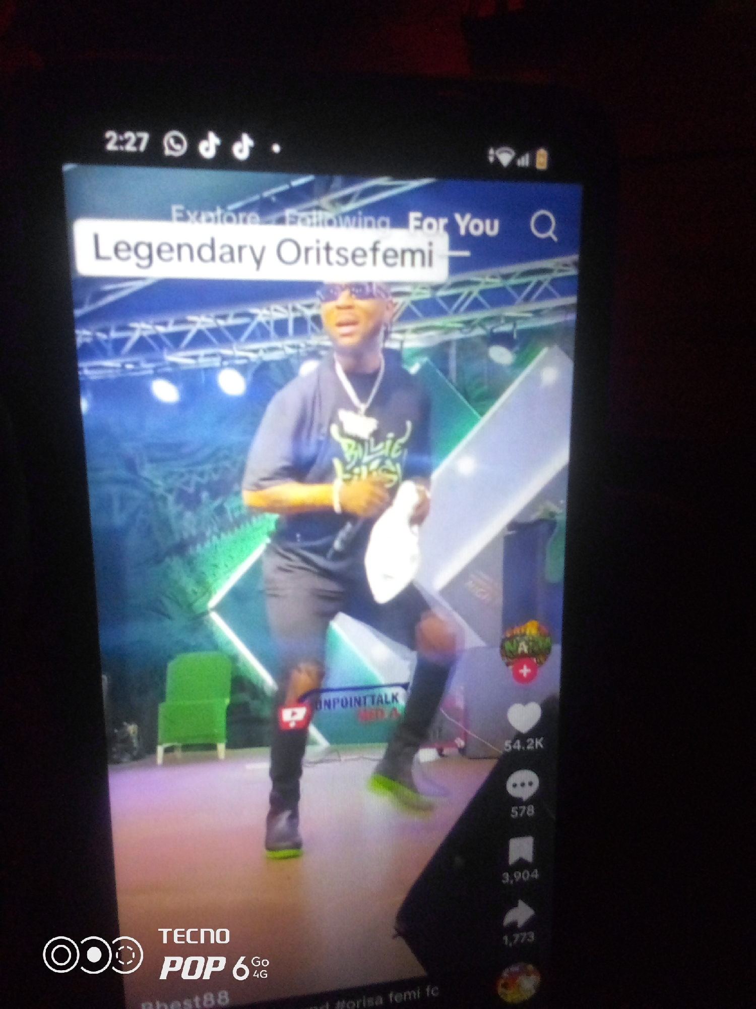 The image shows a screenshot of a mobile device displaying a video with the title "Legendary Oritsefemi". The video appears to be a performance, with a man on a stage wearing a dark t-shirt, black shorts, and sneakers with green soles. He is holding a microphone in his right hand and a white object, possibly a towel or cloth, in his left. The stage has a backdrop with geometric patterns and lighting effects. Social media engagement metrics are visible on the right side of the screen, including likes (54.2K), comments (578), and shares (3,904). A TECNO POP 6 Go 4G logo is present in the bottom left corner, indicating the device used to capture the image. The location of the city of Howayi, Nigeria is not explicitly mentioned or visually represented in the image. The scene is a digital capture of a video being viewed on a phone, not a direct photograph of a physical location.