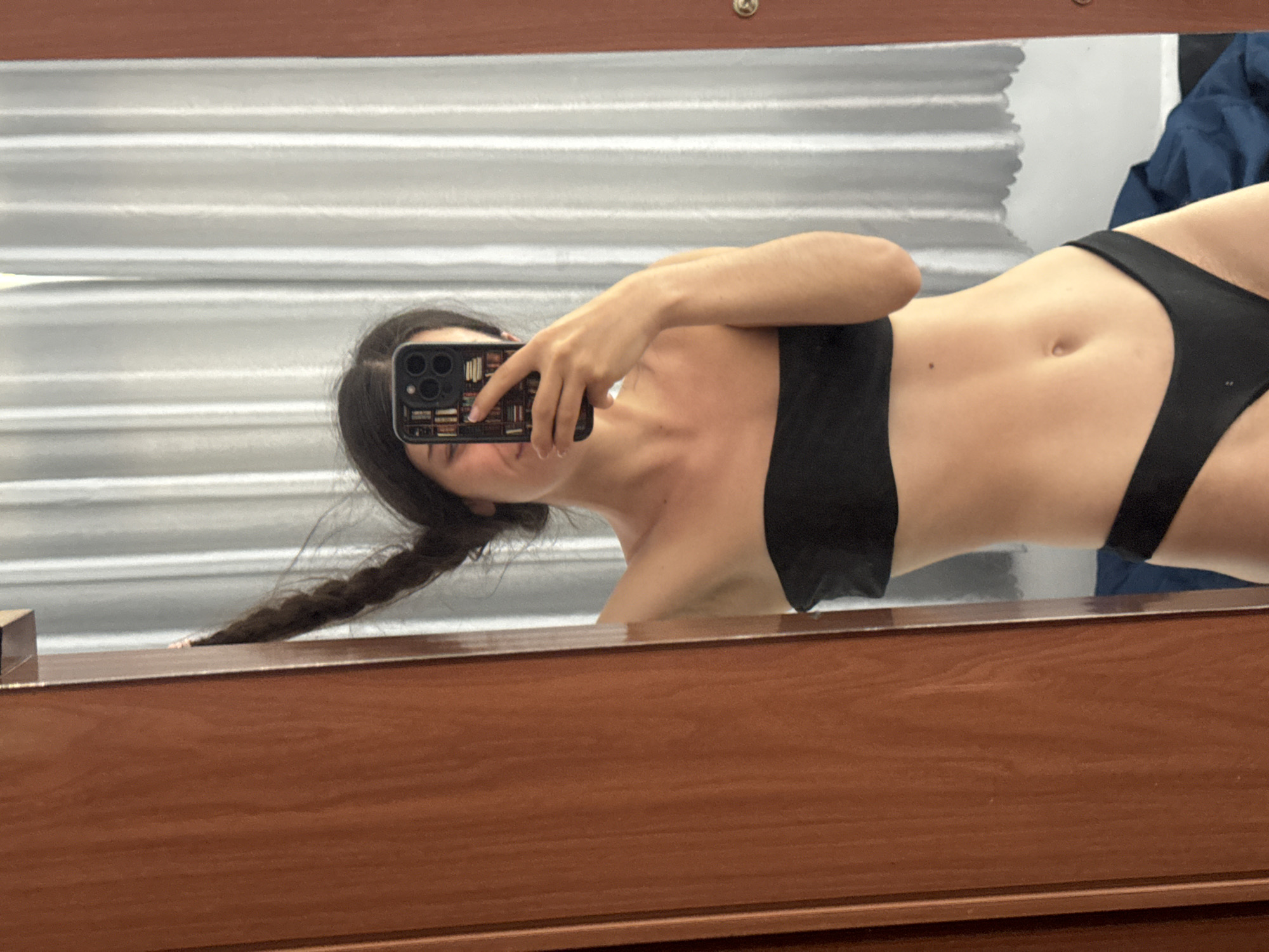 An eye-level mirror selfie captures a person in an indoor setting in Mũi Né, Vietnam. The individual, appearing to be a young woman, is positioned horizontally, seemingly reclining or lying down, with her head tilted to the right. She is wearing a black bikini top and matching black bikini bottoms, showcasing her midriff. Her long, dark hair is styled in a braid that extends towards the bottom left of the frame.
Her face is largely obscured by the smartphone she holds to take the photo. The phone features a prominent multi-lens camera setup on its rear and is encased in a distinctive case with a pattern resembling a bookshelf filled with books.
The selfie reflects a room with a light-colored, possibly white, wall featuring horizontal slats or blinds in the background. A dark blue or black item, possibly clothing or a bag, is visible in the upper right background corner. The lower portion of the image is dominated by the reflection of a dark brown wooden surface, likely a dresser or vanity, on which the mirror rests. The overall impression is one of a casual, personal moment captured indoors, potentially in a hotel room or changing area, consistent with a beach resort location like Mũi Né. No explicit text is visible, though the phone case features a detailed graphic.