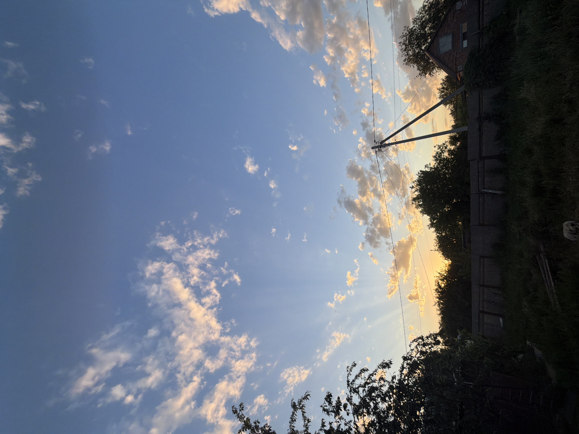 This media file captures a skyward view, likely from a rural or suburban setting in Leningradskaya, Russia. The primary subjects are the sky, filled with scattered white and gold-tinged clouds, and the elements of a human-made landscape encroaching from the right side of the frame. These include a weathered brick building with a boarded-up window, a gray fence, a utility pole with visible wires, and the dark silhouettes of trees and dense vegetation. The lighting suggests it is either dawn or dusk, with the sun casting warm rays through the clouds, creating a serene and somewhat dramatic atmosphere. The presence of a small, light-colored animal, possibly a dog, is barely visible in the upper right quadrant, adding a touch of life to the scene. No text is visible. The overall mood is peaceful, perhaps evoking a sense of quiet contemplation or the end of a day.