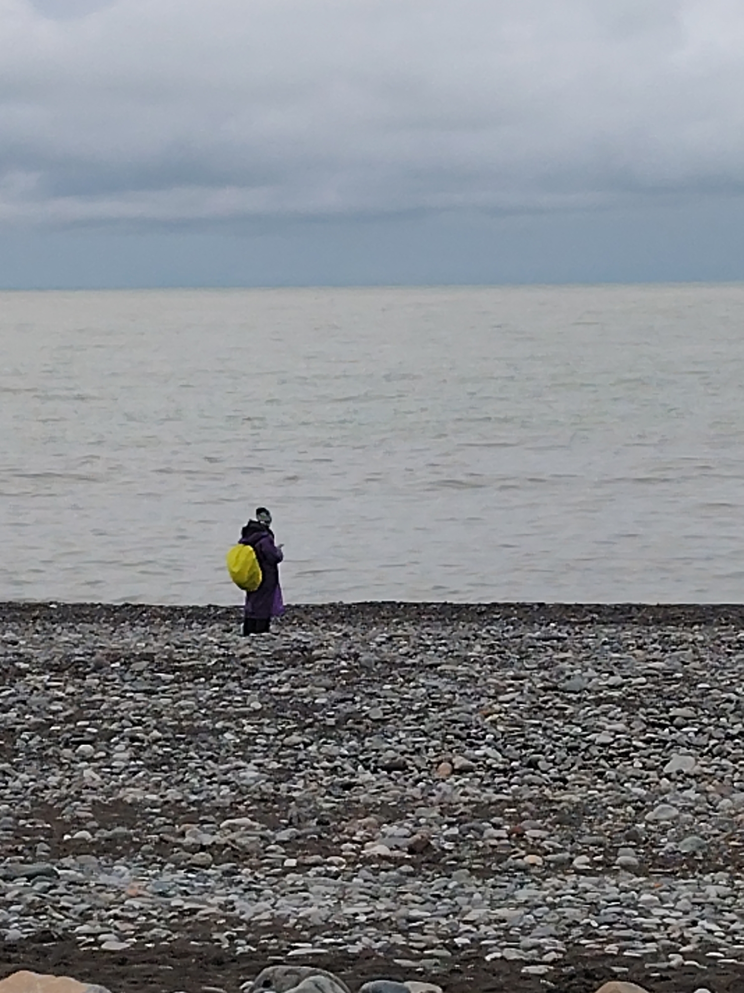 A person stands on a pebble beach in Batumi, Georgia. The person is wearing a purple coat and carrying a bright yellow backpack. The sea is calm and grey, with a cloudy sky overhead. The beach is composed of small, grey pebbles. The scene is devoid of other people or objects.
