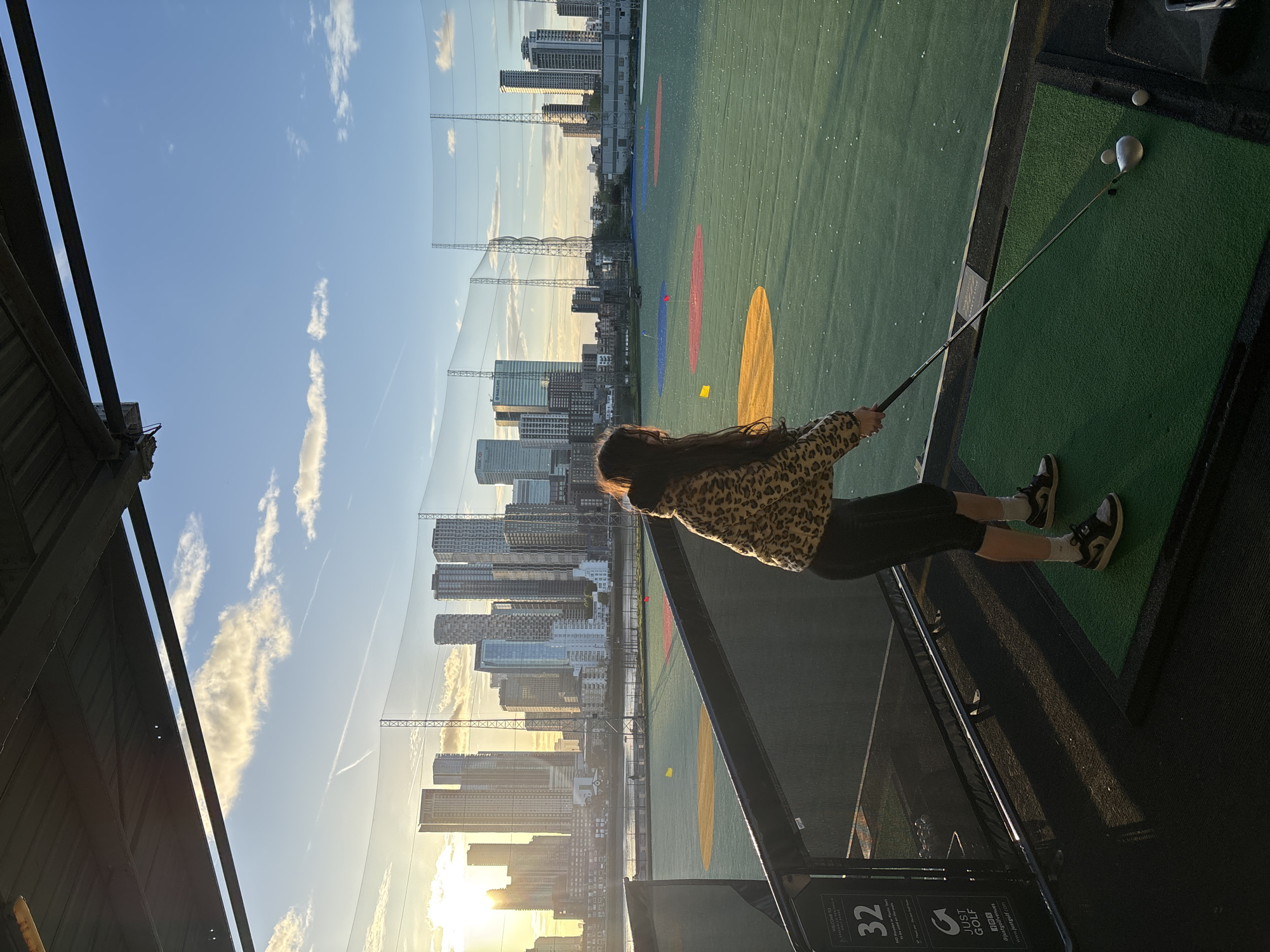 The image depicts an outdoor golf driving range at what appears to be either sunrise or sunset, characterized by warm, golden light casting long shadows across the scene. A woman, viewed from behind and slightly to the right, is the central figure. She has long dark hair and is dressed in a leopard print jacket, dark trousers, white socks, and sneakers. She is actively engaged in golf practice, holding a golf club – specifically a driver or wood – poised above a golf ball on a tee, ready for a swing.
The driving range itself features artificial green turf with several colored circular targets (red, yellow, and blue) scattered across the field. Behind the range, a series of tall, translucent nets extend into the distance, designed to catch golf balls. Beyond these nets, a dense, modern cityscape unfolds, comprising numerous high-rise buildings of contemporary architectural styles. The low sun illuminates the city, creating striking silhouettes and reflections. The sky is a clear blue with scattered white clouds, some exhibiting faint contrails.
In the foreground, to the right, a black panel is visible, displaying the number "32," which likely denotes the golf bay. Other text on this panel includes the "JUST GOLF" logo, the website "www.justgolf.com," and a "BBC" logo. Faintly visible on one of the distant buildings within the cityscape is an "HSBC" logo, a detail consistent with London, United Kingdom, which is the provided location context. The overall setting combines urban modernity with a recreational sports activity, captured during a picturesque time of day.