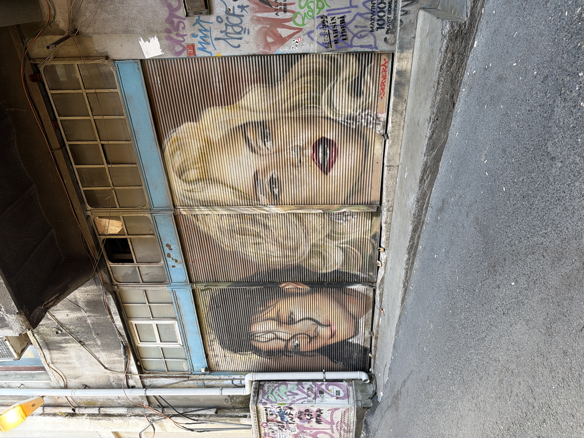 The image depicts a narrow, paved street in Beyoğlu, Turkey, likely an alleyway, with a slightly upward slope. On the left side of the street is a grey utility box covered in colorful graffiti. Adjacent to it, and continuing across the scene, is the facade of a building. The building's ground floor features two large, corrugated metal roll-up doors, each painted with a mural of a woman's face. The left door shows the face of a woman with dark hair and piercing eyes, her features partially obscured by dark, wavy lines. The right door displays the face of a blonde woman with striking red lips and blue eyes. The windows above the roll-up doors are framed in blue metal and contain multiple panes of dusty glass. The upper portion of the building is constructed from rough, grey concrete, with exposed electrical wires and conduits running across its surface. A yellow street lamp is visible at the top left corner of the image, extending from the building. The pavement in front of the building is worn and cracked, with a raised concrete curb. The street itself is asphalt, littered with numerous cigarette butts. The overall impression is one of urban decay and vibrant street art.