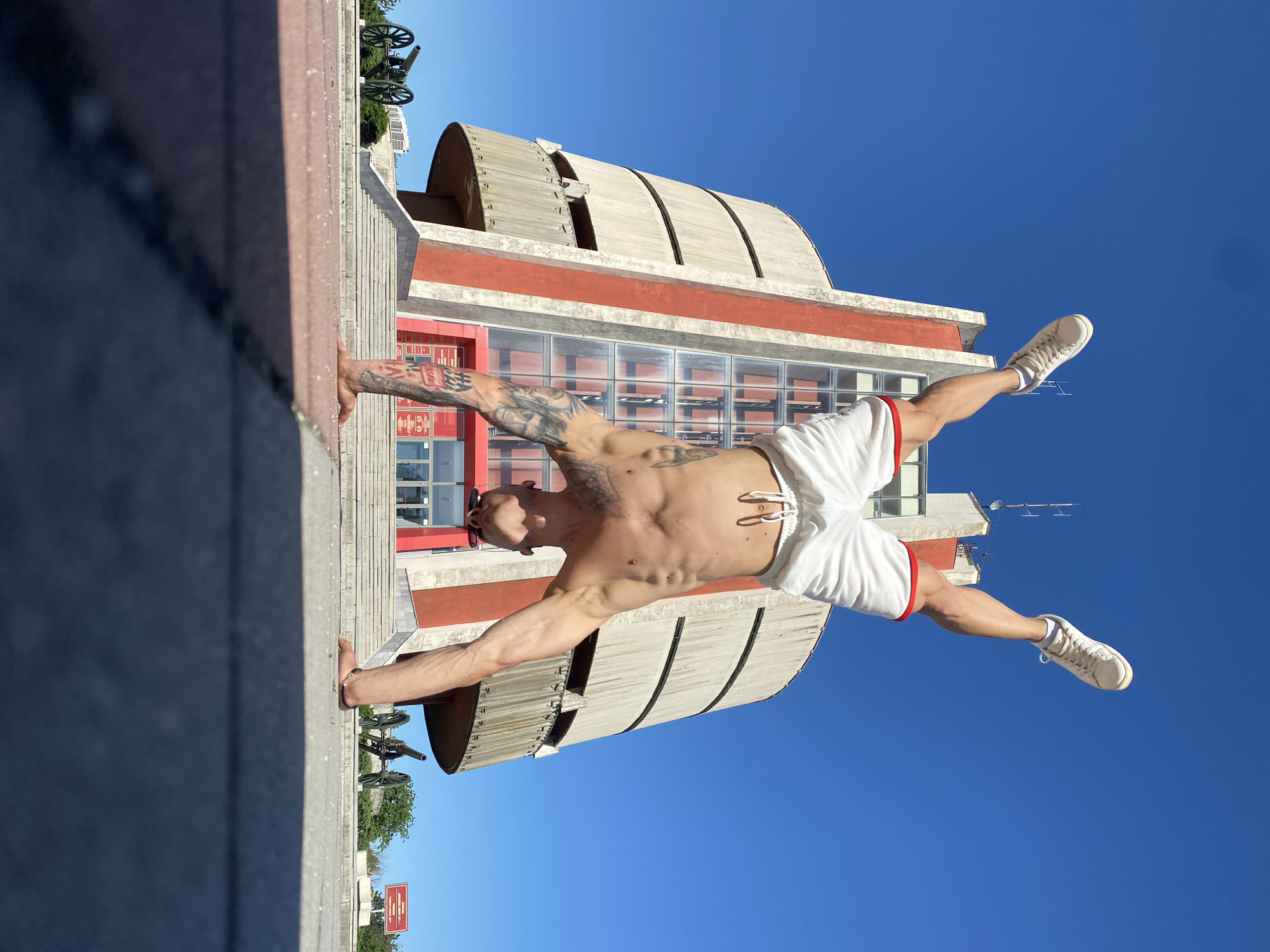 A shirtless man wearing white shorts and white sneakers is performing a handstand outdoors in Pleven, Bulgaria. He is balanced between a grey concrete wall and the steps of a building with a rounded roof. The building is red and white and has many windows. Behind the building, the sky is clear and blue. To the left, there is a cannon on a grey concrete platform. A small sign is in the lower left corner.