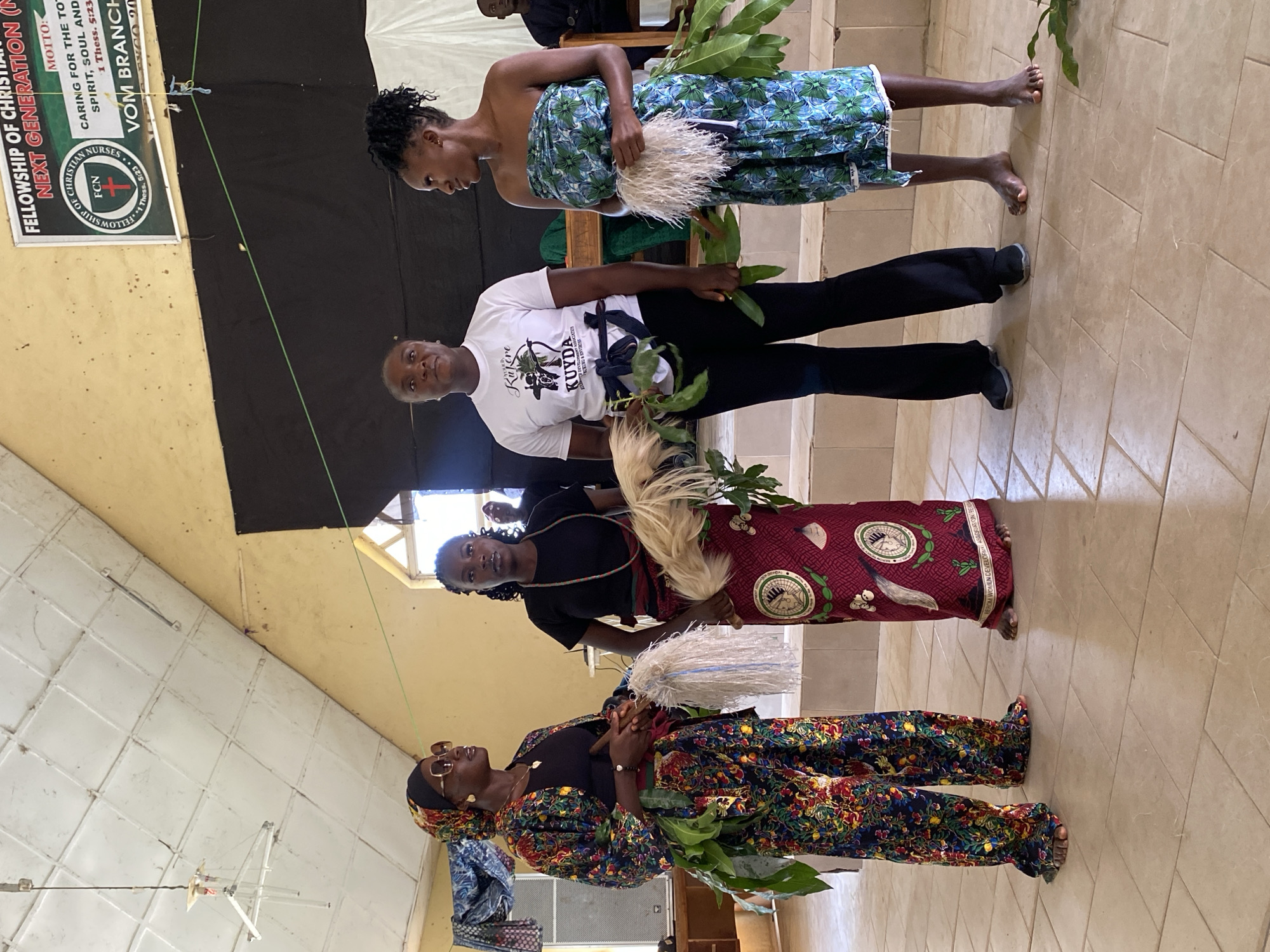 This media file depicts an indoor scene with several individuals, likely at an event in Dutsen Kato, Nigeria. The lighting suggests it could be daytime, with natural light possibly coming from a window or open space.
The foreground features two women, one wearing a vibrantly patterned, traditional outfit and the other in a black top and a dark, patterned skirt. Both are holding leafy branches and appear to be part of a procession or performance. The woman in the patterned outfit is also wearing sunglasses. Behind them, another woman is visible, dressed in a white t-shirt with text and dark leggings, holding similar leafy branches. Further back, a woman in a blue and green patterned wrap is standing.
In the background, a banner is visible with text that includes "FELLOWSHIP OF CHRISTIAN", "NEXT GENERATION", "MOTTO: CARING FOR THE TOTAL SPIRIT, SOUL AND BODY", "VOM BRANCH", and "1 Thess. 5:23". This suggests the event is likely a religious gathering or a community event organized by a Christian fellowship. The overall atmosphere seems celebratory or ceremonial, indicated by the attire and the props. The floor is tiled, and the walls appear to be a light color, possibly painted. The presence of the banner and the traditional attire points towards a cultural and religious context within Nigeria.