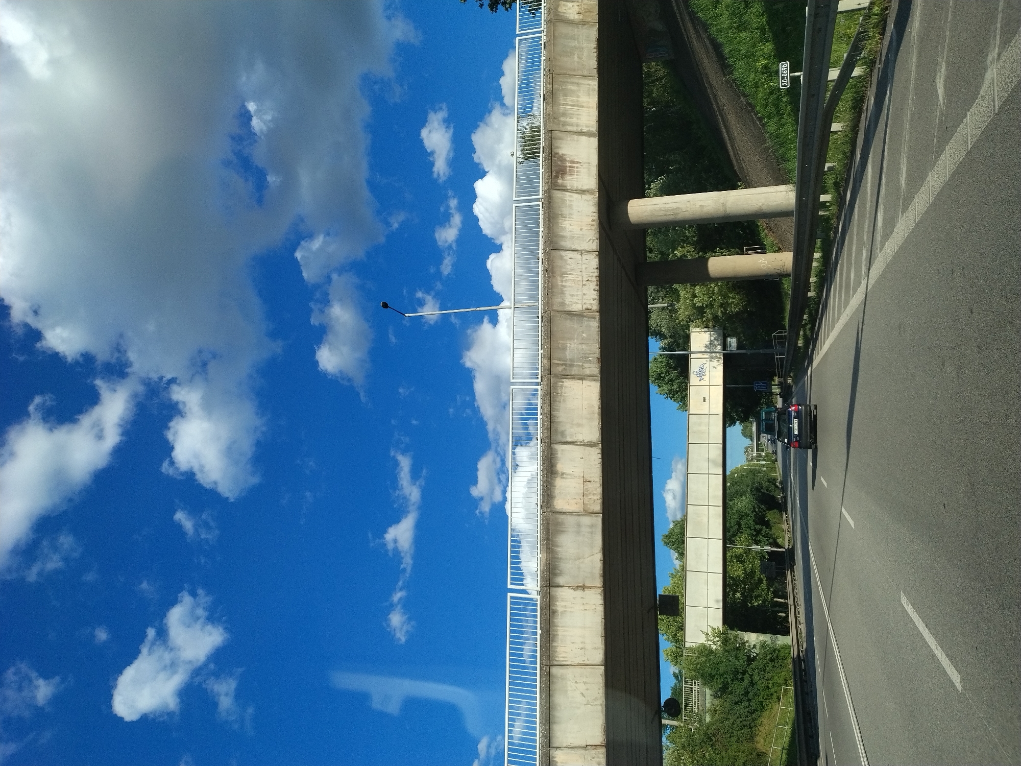 The image depicts a view from a car driving on a road beneath an overpass in Písek, Czechia. The sky is a vibrant blue with scattered white clouds. The overpass, constructed with concrete and featuring a white metal railing, runs horizontally across the upper half of the frame. Below this overpass, another, smaller pedestrian bridge is visible, also with a white railing. The road surface is asphalt, marked with white lines. A dark-colored car is driving ahead in the right lane. Green trees and grass line the sides of the road, with a grassy embankment to the right of the road. A metal guardrail runs along the edge of the embankment. There are signs visible along the road and on the embankment. The lighting suggests it is daytime, possibly late morning or early afternoon, given the shadows.