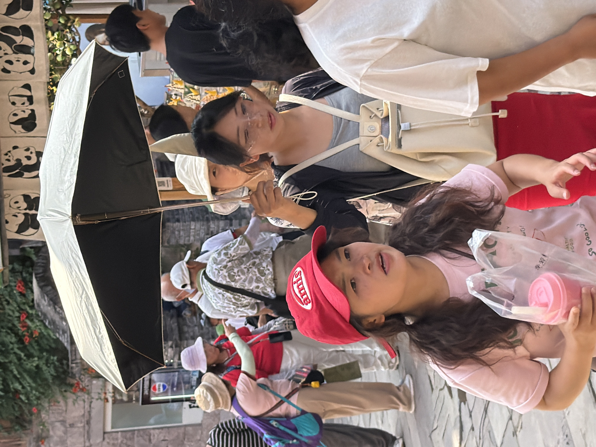 In Yuzhong District, China, a diverse group of people are gathered under a large black and white umbrella. In the foreground, a young woman wearing a red baseball cap and a pink t-shirt holds two plastic bags containing drinks, one of which is a pink beverage. To her right, a person in a white t-shirt stands with their arm extended. Behind them, a woman with glasses wears a black jacket over a grey tank top and a light-colored backpack. Several other individuals, some obscured by the umbrella and the crowd, are visible in the background. The environment appears to be an outdoor market or pedestrian street, with stone paving and buildings suggesting an urban setting. Decorative banners featuring pandas hang above.