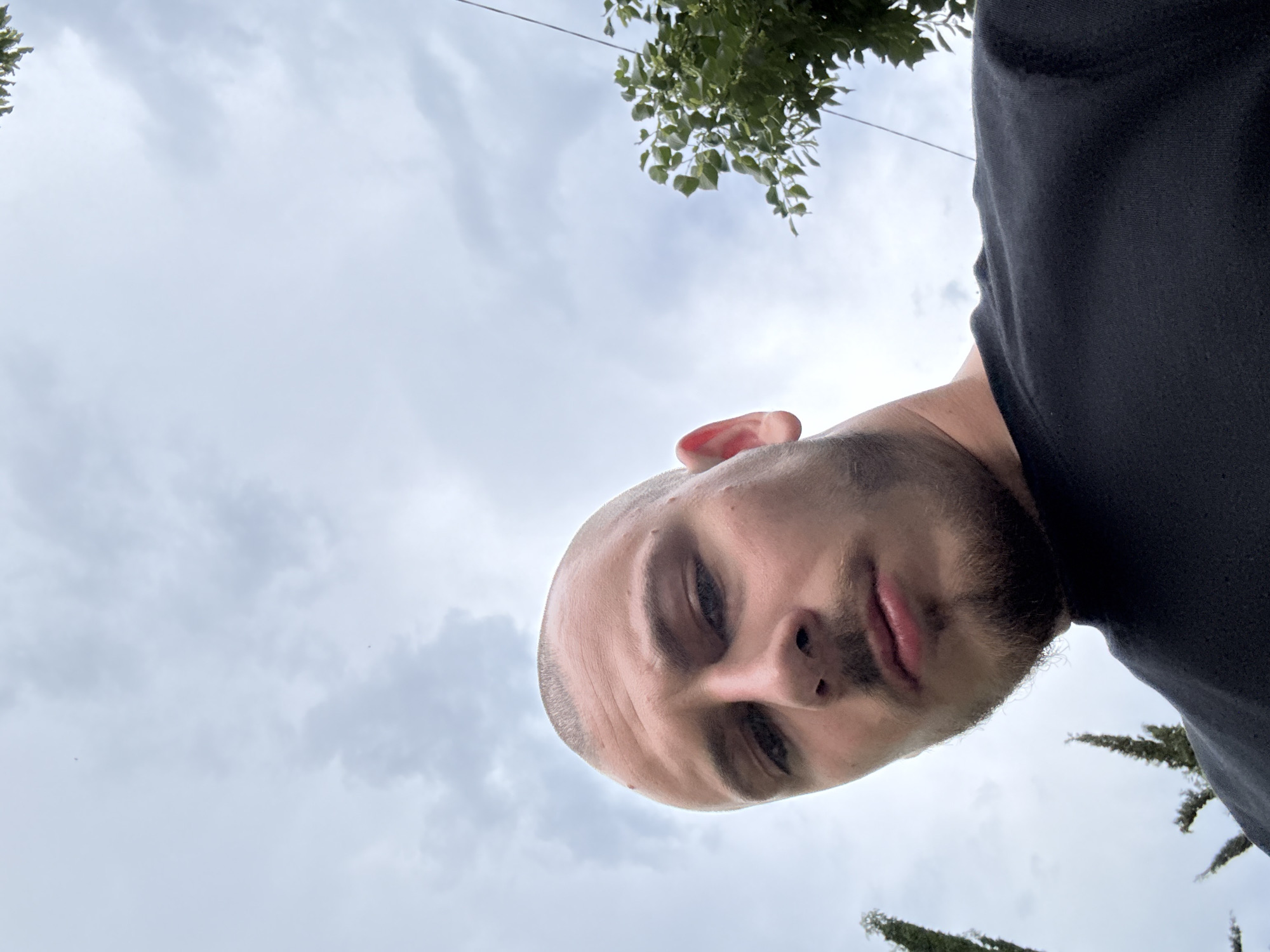 The photograph presents a low-angle, close-up view of a bald man with fair skin, a short beard, and facial hair. The man is wearing a black shirt. Above him, a cloudy sky is visible, with glimpses of green foliage in the upper corners. The photograph appears to be taken outdoors. The location is not discernible from the image itself and there is no indication that this image was taken in Stara Zagora, Bulgaria.