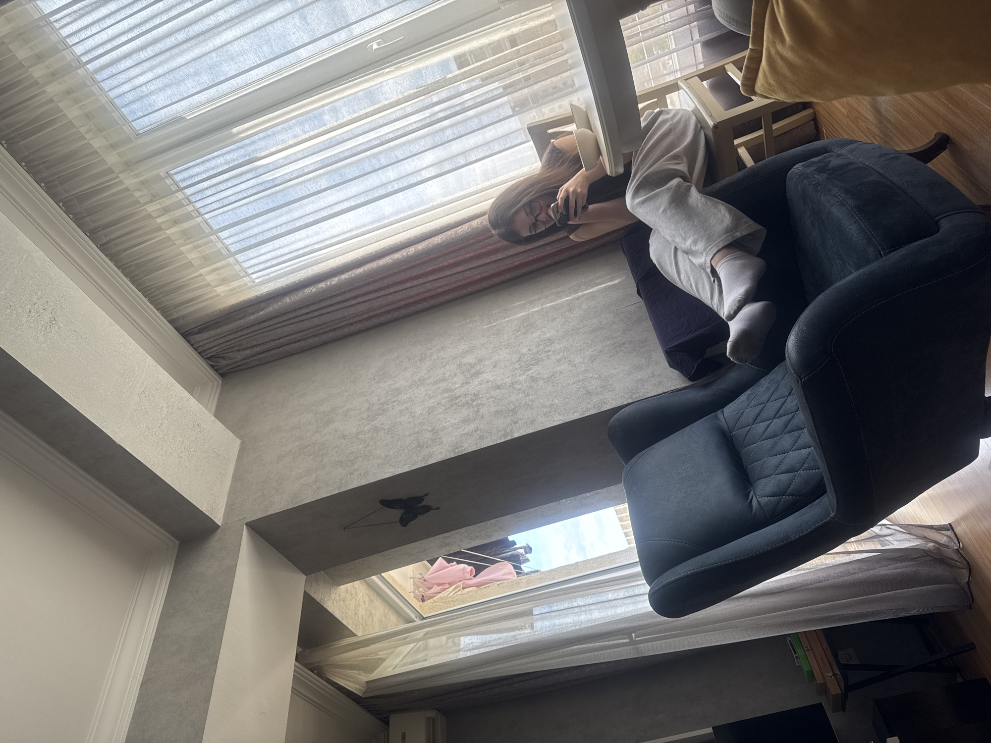 A person is sitting in an armchair facing a window, holding a phone, presumably taking a selfie. The room has several windows allowing ample light. The walls are painted in neutral tones, and there are some decorative elements present. Given the architectural style, the location appears to be in Tbilisi, Georgia, a city known for its distinct urban landscape.