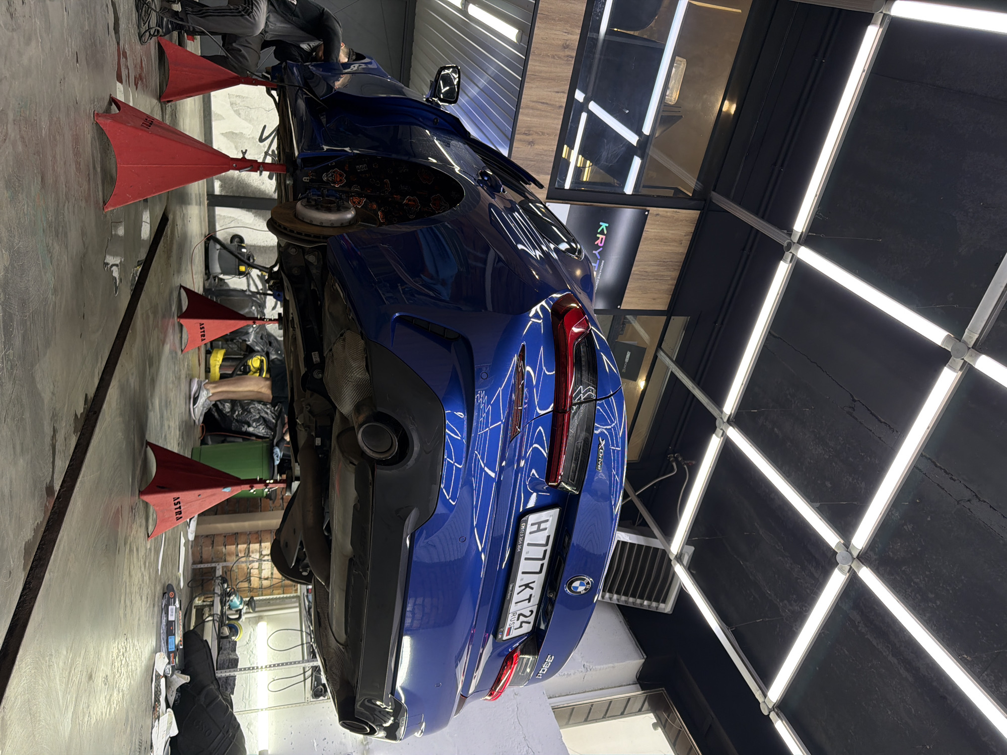 A blue BMW X-Drive, model 330d, is suspended on red Astra jack stands in what appears to be an auto shop. The car has the Russian license plate H777 KT 24, indicating it is located in the city of Krasnoyarsk, Russia. The underside of the car is visible, and the presence of people suggests that maintenance or repair work is being performed. The shop has black ceilings with square patterns formed by white light fixtures.