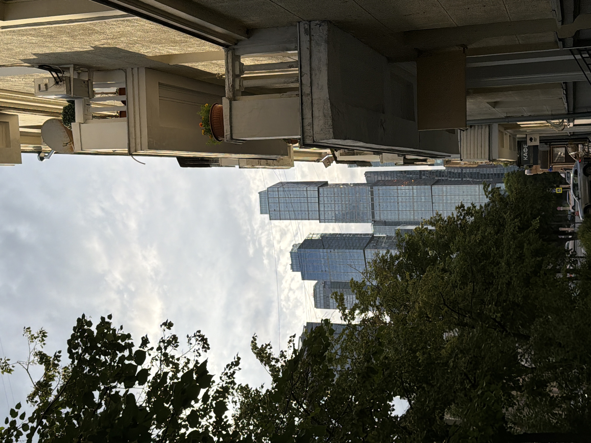 The image depicts a street scene in Moscow, Russia, with modern skyscrapers in the background juxtaposed against an older building in the foreground. The older building, on the right side of the frame, has a textured facade and balconies adorned with plants. One balcony features a satellite dish and an air conditioning unit. Below, street-level businesses are visible, including one with a sign displaying "КАФЕ" (Cafe) in Cyrillic script. Trees with lush green foliage line the street, partially obscuring the view of the skyscrapers. Several cars are parked and driving on the street. The sky is overcast with scattered clouds, and the sunlight casts shadows on the building.