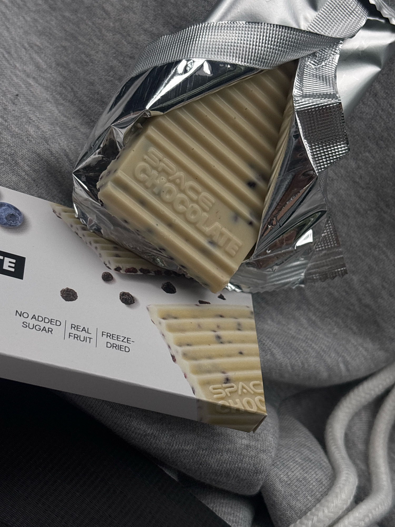 The image displays a white chocolate bar with blueberries and freeze-dried fruit pieces, partially unwrapped from its silver foil packaging. The chocolate bar is ribbed and appears to be a single serving. It rests on a grey, textured surface, likely fabric, possibly a hoodie. Part of the product's white packaging is visible at the bottom of the frame, with text that includes "NO ADDED SUGAR", "REAL FRUIT", and "FREEZE-DRIED". The location in the city of Tsutsyliv, Ukraine is not depicted or suggested by any elements in the image.
