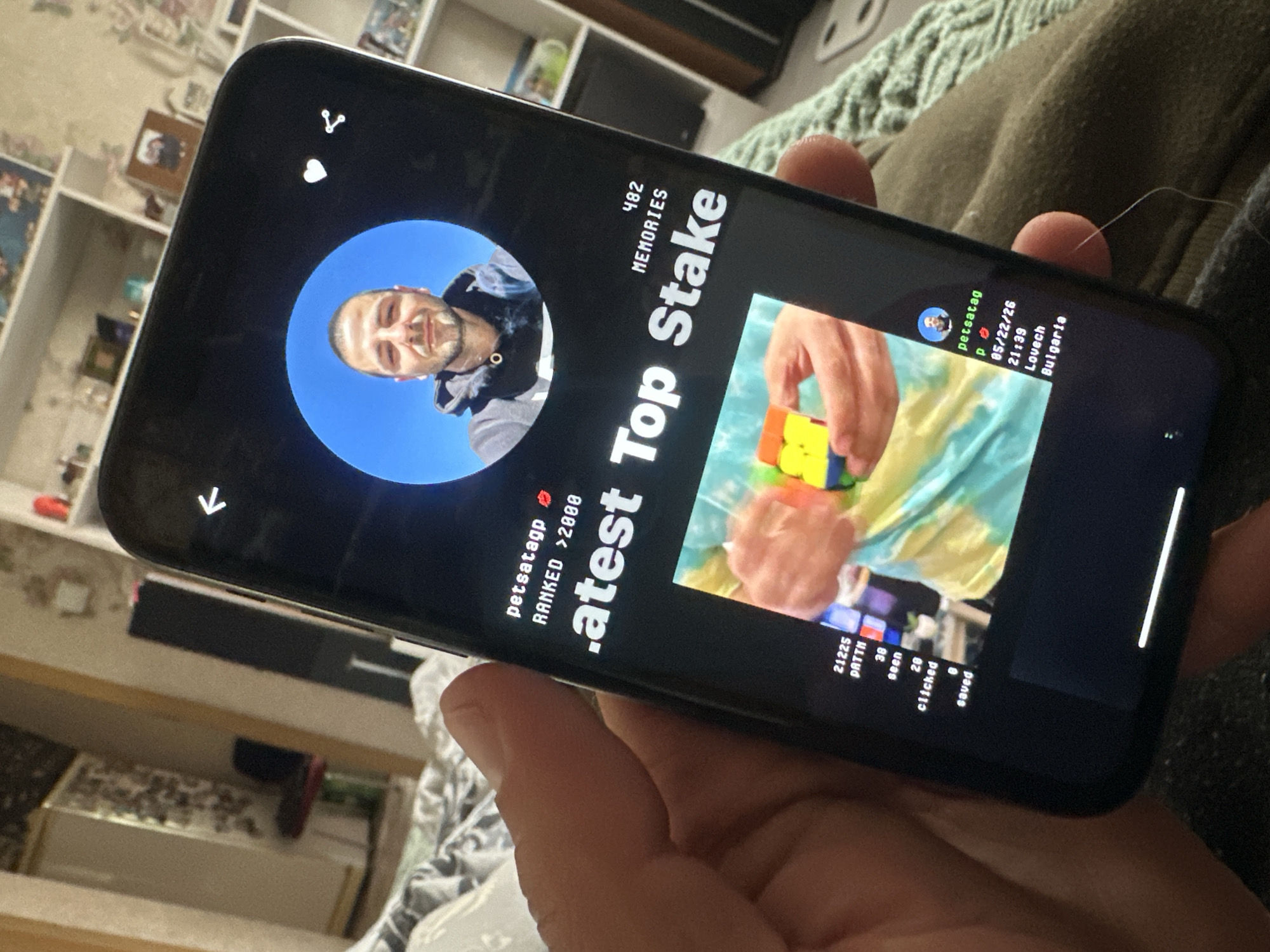A person is holding a smartphone displaying an app with an image of a man and a Rubik's Cube. The app interface shows the username "petsatagp", a ranking ">2000," the number of memories "482," and the text "Latest Top Stake." The location listed on the phone is Lovech, Bulgaria, not Slavyani. The date and time are 05/22/26 and 21:39, respectively. The app also displays metrics such as "21225 PNTTM," "38 seen," "20 clicked," and "0 saved."
