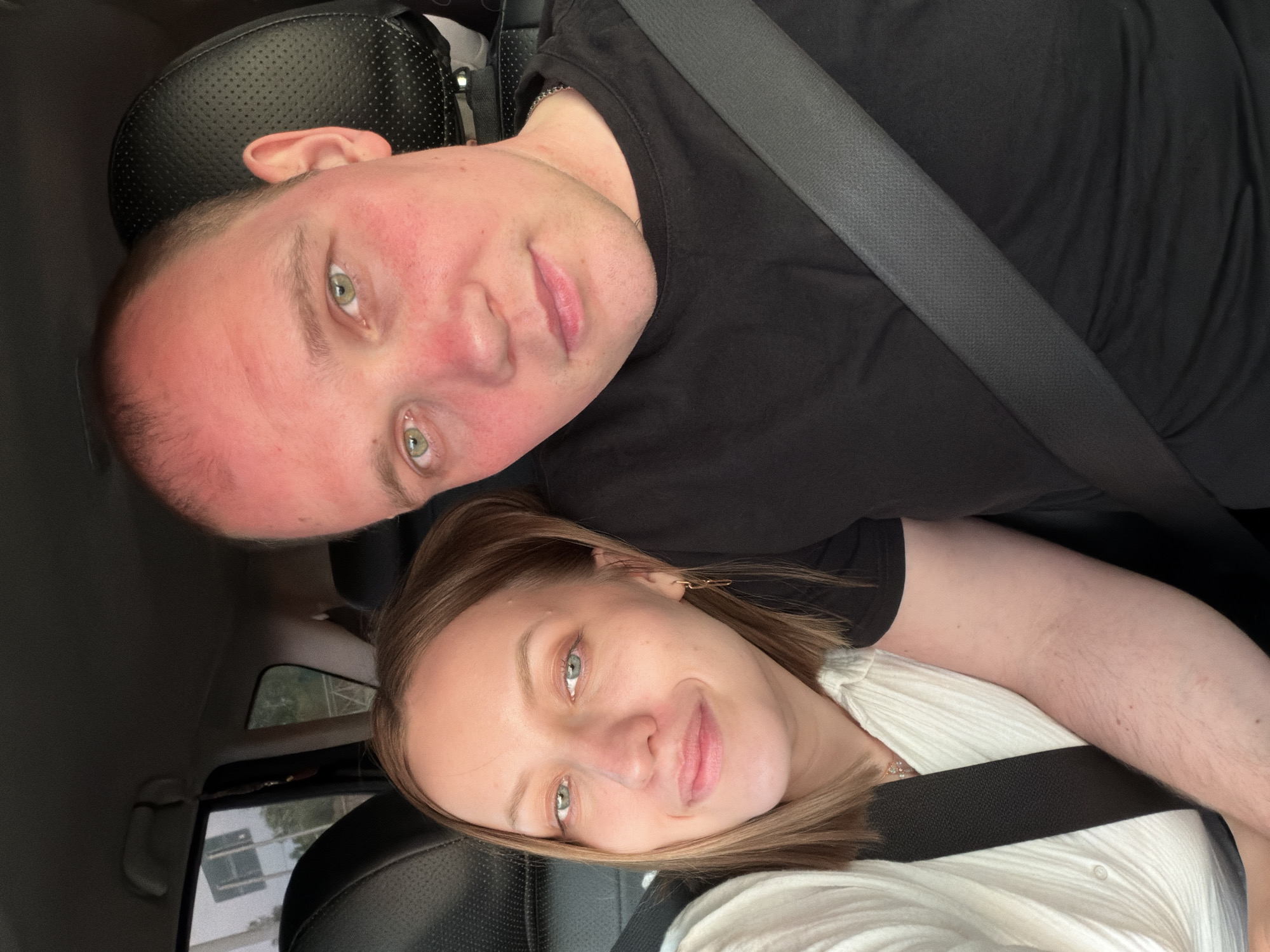 A selfie shows two light-skinned individuals in a car. The male is positioned above, wearing a black shirt and a seatbelt. His face appears flushed. The female is below him, wearing a white shirt and a seatbelt. The interior of the car is visible, including the dashboard and seats. The photo's location is not determinable.