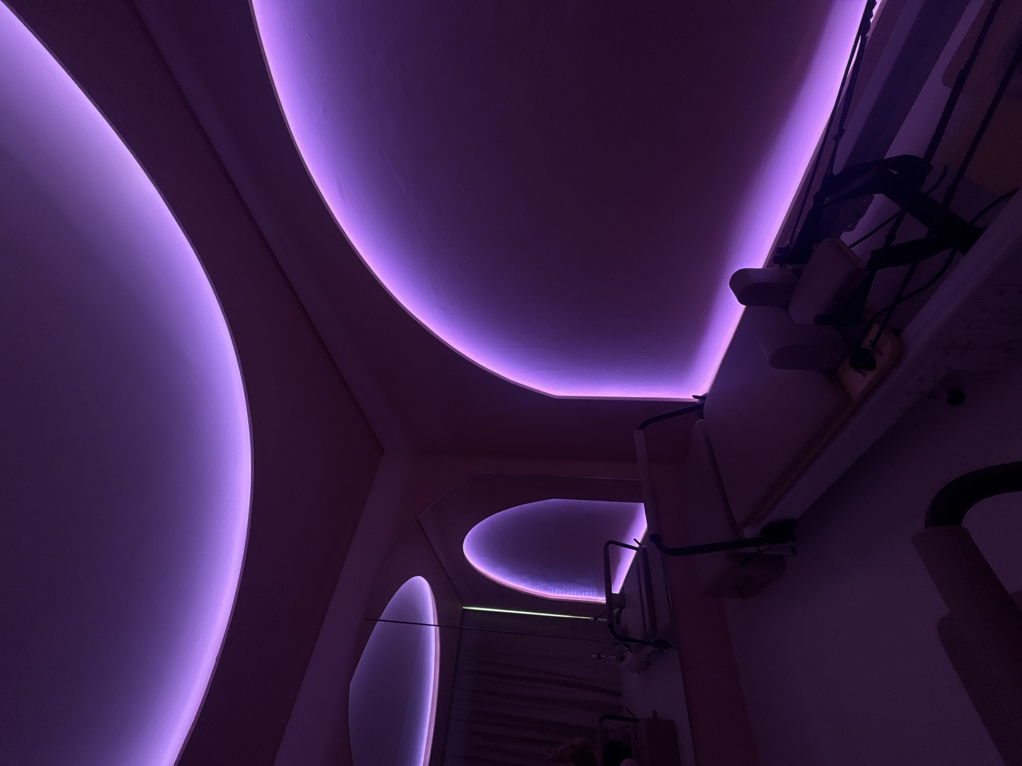 A low-angle interior shot in Mexico City, Mexico, showcases a pink and purple space with curved ceiling elements illuminated by integrated LED lighting. The foreground features a large, rounded ceiling feature with a soft purple glow, juxtaposed against another curved element further back. Architectural details, including pipes and equipment, are visible against a pale pink wall on the right. A smaller, elongated ceiling curve with LED lighting is visible in the background, hinting at a corridor. The overall lighting casts strong shadows and highlights the architectural forms.