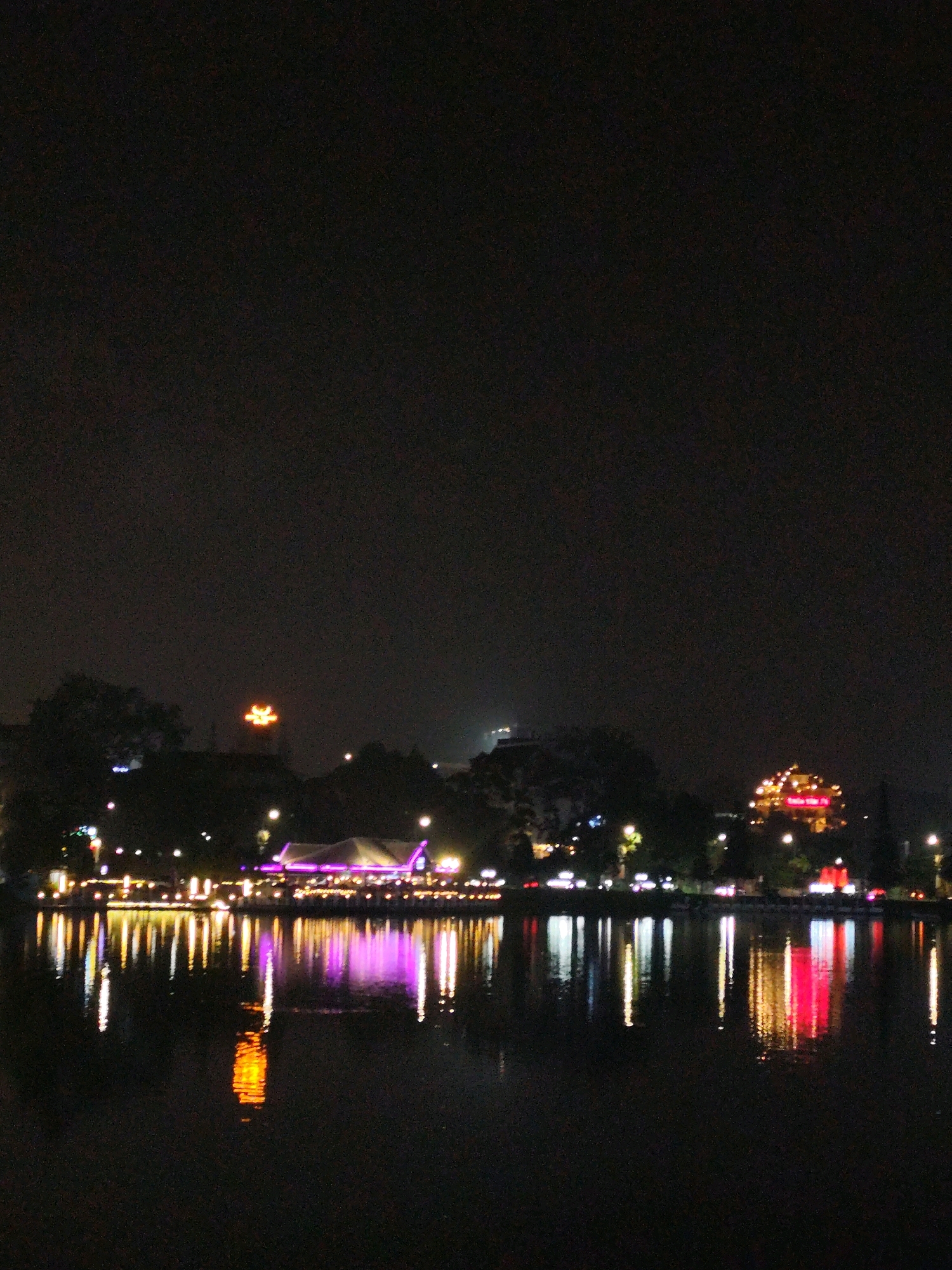 The image depicts a nighttime scene in Da Lat, Vietnam, focusing on the Xuan Huong Lake and its illuminated surroundings. The foreground is dominated by the dark water of the lake, reflecting the lights from the shore. A prominent orange, stylized dragon-like figure is visible in the sky above the buildings. Along the lake's edge, various structures are lit up, including a building with a purple illuminated roof, and a string of lights that appear to be from restaurants or shops. Further in the background, a large, round building is brightly lit with yellow and red lights, possibly a pagoda or a prominent landmark. The overall atmosphere is one of a vibrant, illuminated cityscape at night, with the reflections on the water adding depth and color to the scene. There are no people clearly visible in the image.