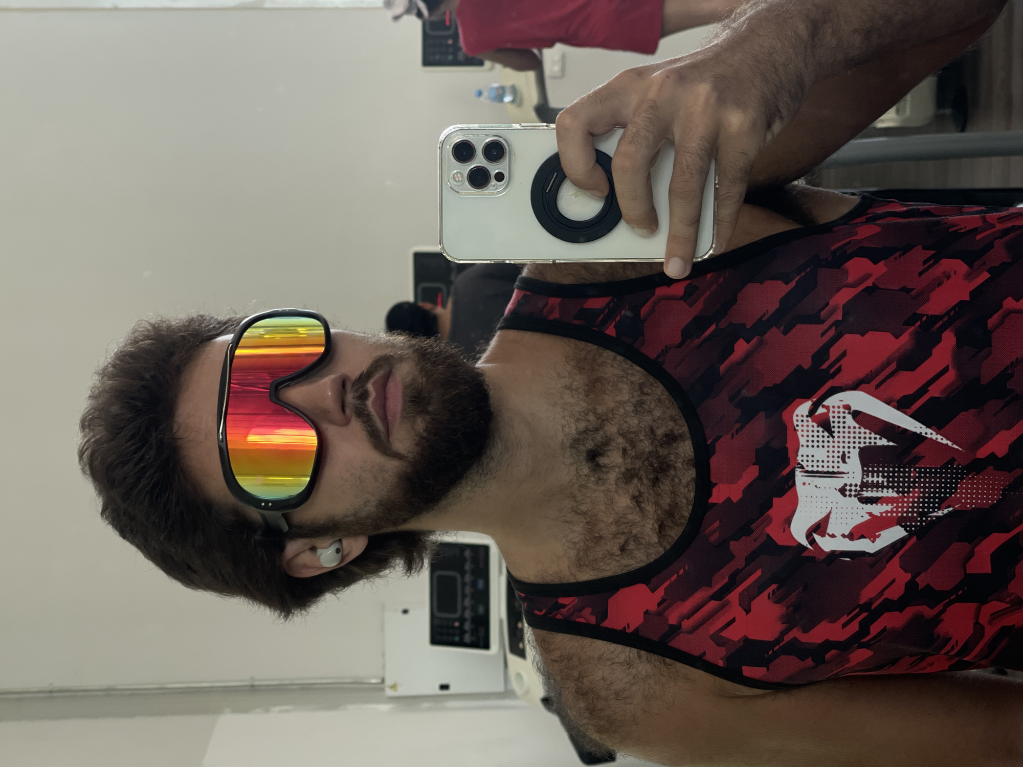 A man is taking a mirror selfie in a gym. He is wearing large, multi-colored mirrored sunglasses and a red and black camouflage tank top. He has a beard and is wearing a white wireless earbud in his left ear. He is holding up a white iPhone with a black ring holder attached to the back. The iPhone camera is pointed towards the mirror, capturing his reflection. In the background, there are reflections of gym equipment, including what appear to be weight machines and a water cooler. There is also a reflection of another person in a red shirt in the background. The lighting suggests it is daytime. The setting appears to be an indoor gym, possibly in Bangkok, Thailand, given the context. The man's expression is neutral. There is no visible text in the image.