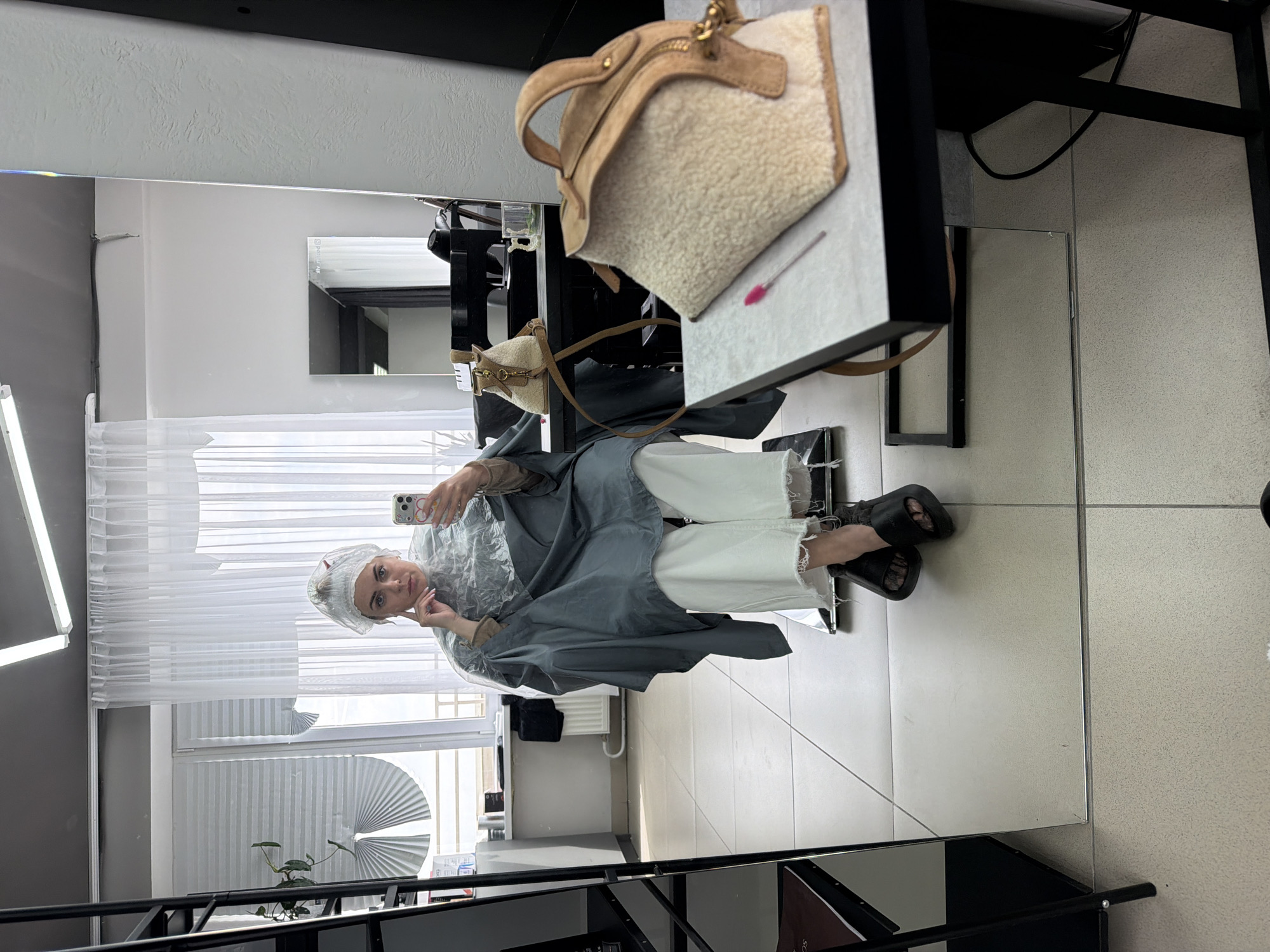 A woman wearing a shower cap and a grey salon cape is seated in a salon chair, holding a smartphone and taking a selfie in a mirror. She is wearing white pants and black platform sandals. The mirror reflects the salon interior, which includes white curtains, a window with a pleated shade, a plant, and a white-tiled floor. In the foreground, a handbag sits on a mirrored surface. The location is Votkinsk, Russia.