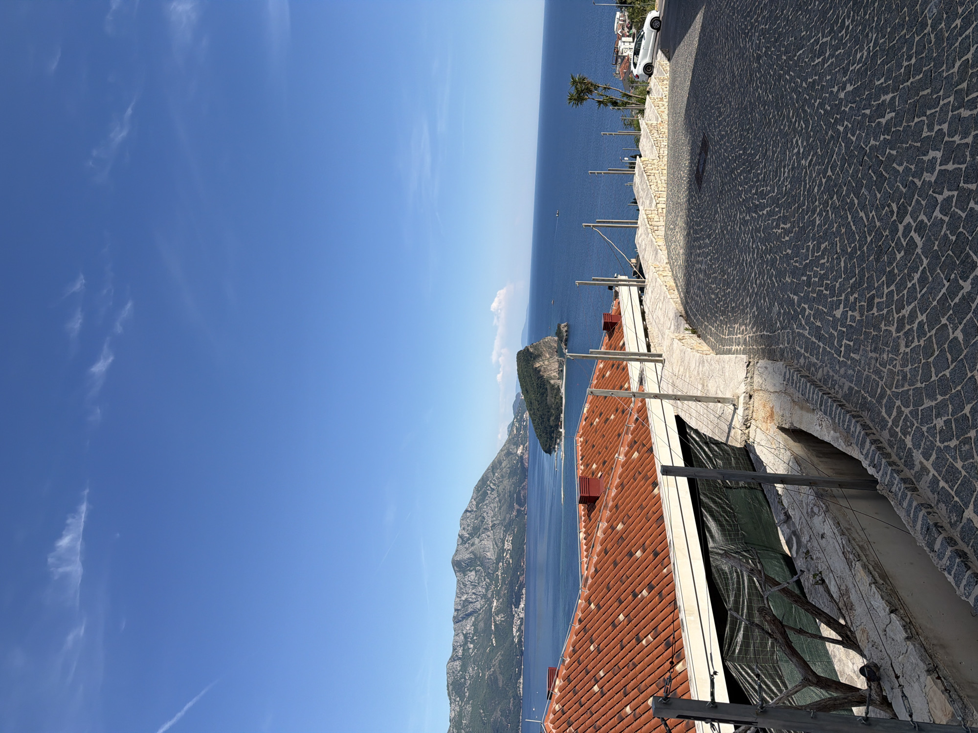 This image presents a vibrant, sun-drenched coastal landscape in Budva, Montenegro, captured from an elevated perspective looking down towards the sea. The scene is dominated by a clear, expansive blue sky with a few wispy white clouds, meeting a deep blue, calm sea at the horizon.
In the mid-ground, rugged mountains and cliffs, partially covered in green vegetation, stretch along the coastline, creating a dramatic backdrop to the azure waters. A small, green-topped rocky island or peninsula is visible in the sea. In the distance, tiny specks suggest boats or ships on the water.
In the foreground and to the right, a curving cobblestone road or path is visible, showing distinct shadows that indicate bright sunlight. A white car is parked on this road further up the incline. Below the road, residential buildings with characteristic orange/terracotta tiled roofs descend towards the sea. Stone walls, supports, and green tarps covering lower areas are also visible, suggesting architectural elements common to the region. A palm tree adds to the Mediterranean feel of the setting.
The photograph captures a tranquil, scenic moment under clear, sunny weather conditions, likely during midday or early afternoon. No people are visible, and no specific activity is taking place. There is no visible text in the image.