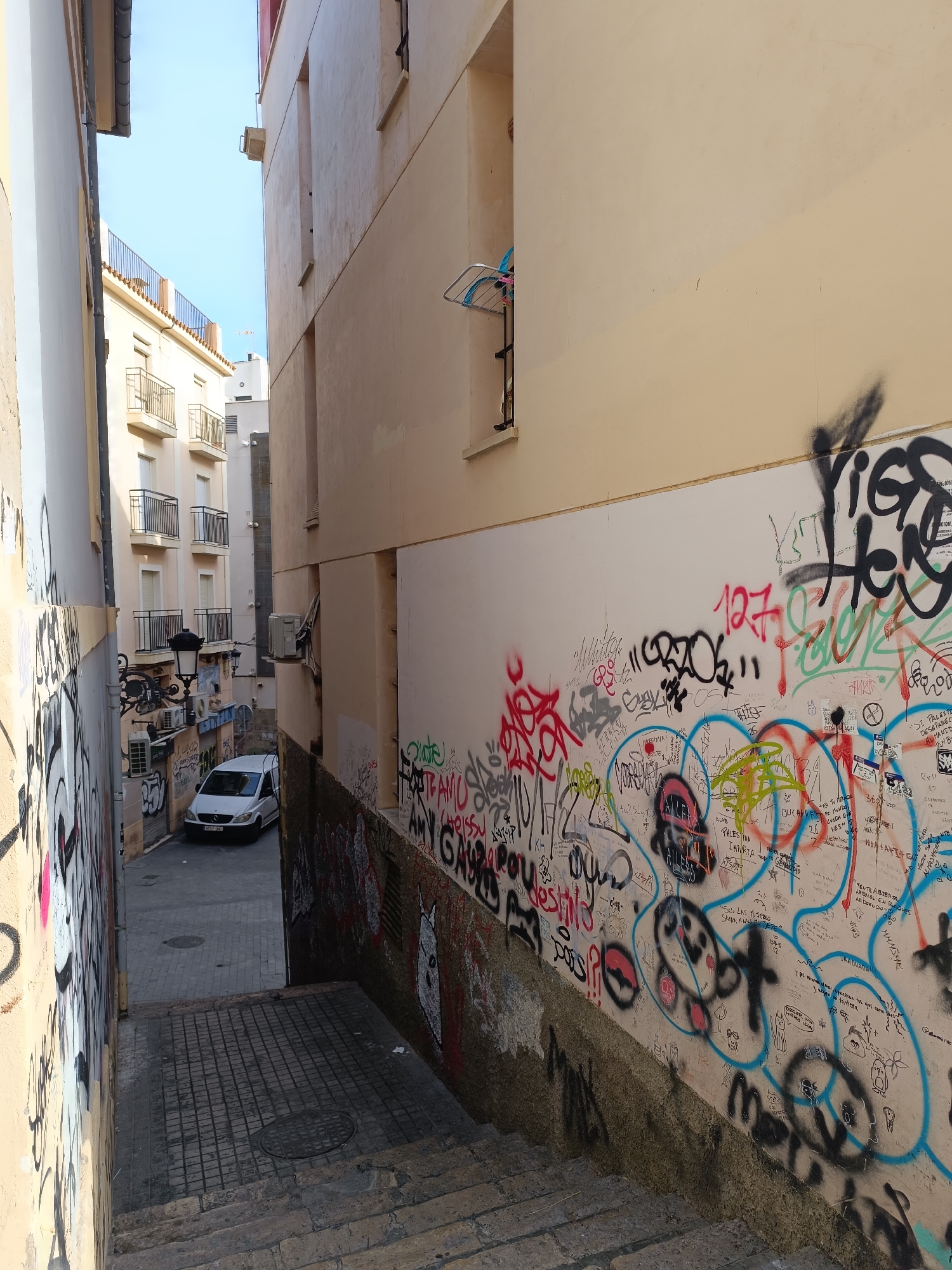 An upward-angled, medium shot captures a narrow alleyway with tall, beige-colored buildings on either side. The alley is filled with graffiti. The left building is partially visible, its wall covered in black and pink graffiti. The right building's wall is also extensively graffitied with various colors and styles of spray paint and markers, including caricatures, lettering, and abstract designs. A white van is parked in the distance at the end of the alley, which appears to be paved with cobblestones. Above the van, a black lamppost with ornate detailing is visible, along with several air conditioning units mounted on the buildings. The sky is a clear, pale blue. In the upper right portion of the image, a clothesline with some laundry is attached to a window frame. The foreground of the image shows stone steps leading down into the alley.