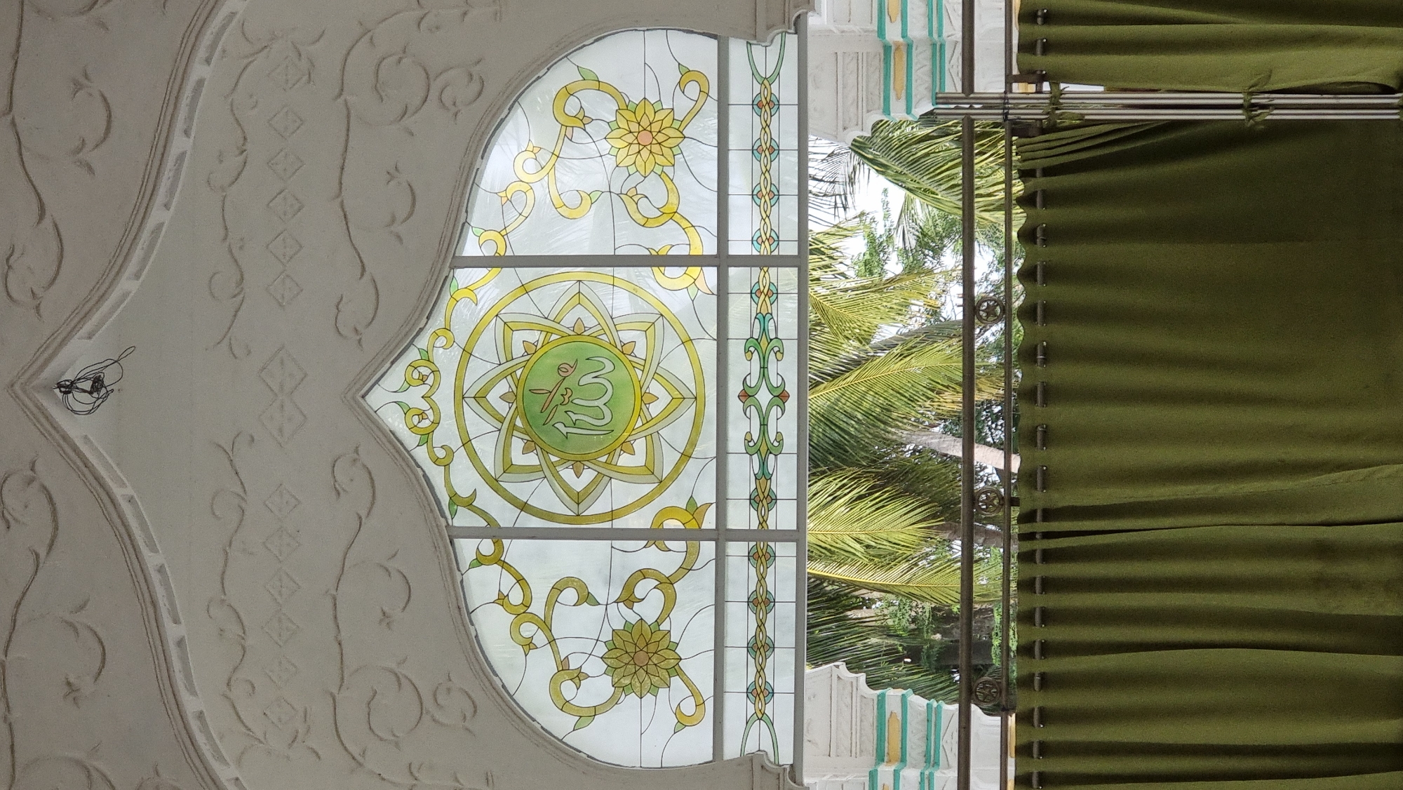 The image shows an arched stained-glass window with Islamic calligraphy in the center, set against a white decorative archway. Below the window, there is a railing with two green curtains hanging in front of it. Palm trees are visible in the background through the window. The scene appears to be inside a building, possibly a mosque or a place of worship, in Lapang Timu, Indonesia. There are no people visible in the image. The lighting suggests it is daytime.