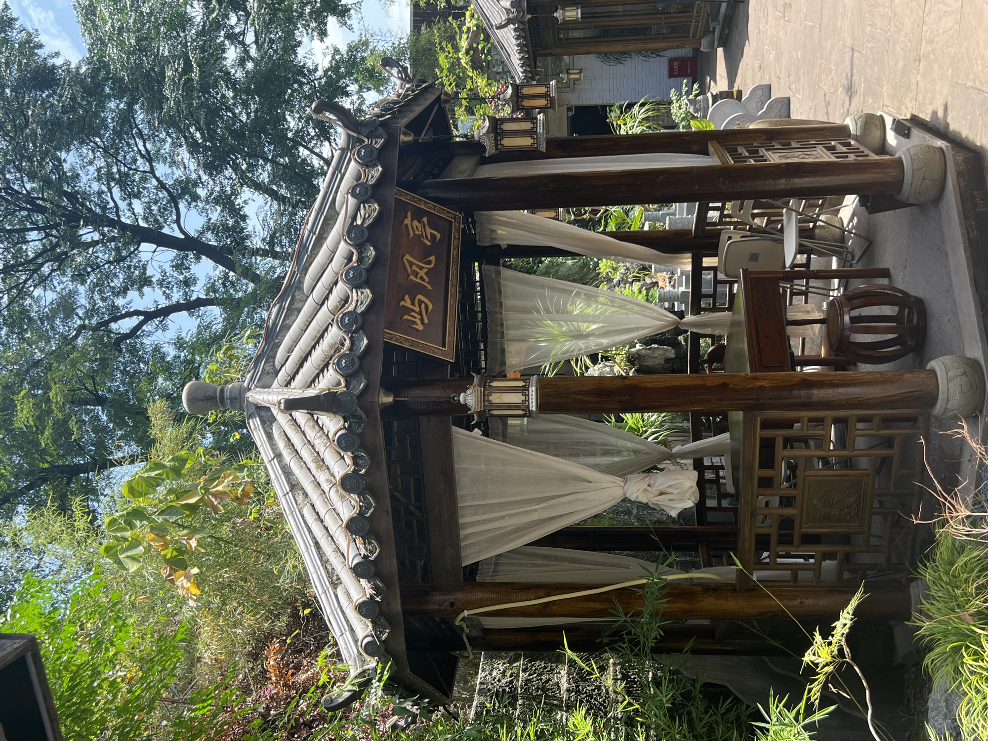 A photograph captures a serene outdoor seating area in Yuzhong District, China, likely a garden or courtyard. The central focus is a traditional wooden gazebo with a grey tiled roof. The gazebo is furnished with a dark wooden table and chairs, some of which are folded. White sheer curtains hang on three sides of the gazebo. Ornate lanterns are suspended from the roof. A wooden sign with Chinese characters is mounted above the entrance. The area is surrounded by lush greenery, including bamboo and other foliage, and the ground is paved with stone. In the background, another structure with a similar architectural style is visible. The scene is illuminated by natural daylight.
