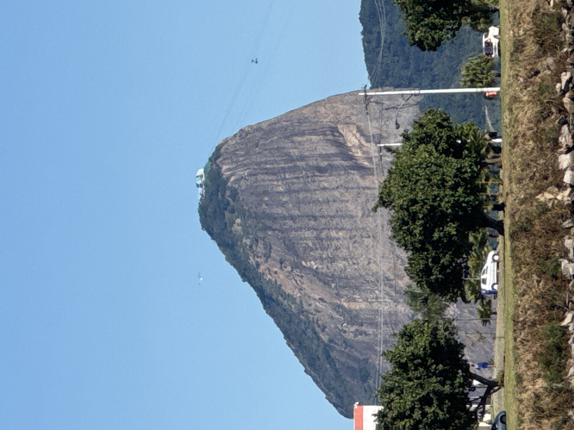 The image shows a rocky mountain with lush green trees in the foreground and a clear blue sky. In the distance, cable cars are visible ascending the mountain, suggesting a tourist attraction or transportation system. A white van with blue accents is parked on a grassy area near the mountain base, and several people are present, some walking and others near the van. A small white structure with red accents is visible at the bottom of the image. The sun is shining brightly, indicating it is daytime, and the weather appears to be clear and pleasant. The overall scene evokes a sense of leisure and outdoor activity in a natural setting. The image appears to be taken from a slightly elevated viewpoint, looking towards the mountain and the surrounding landscape. The presence of cable cars and a well-maintained grassy area suggests a developed tourist destination.