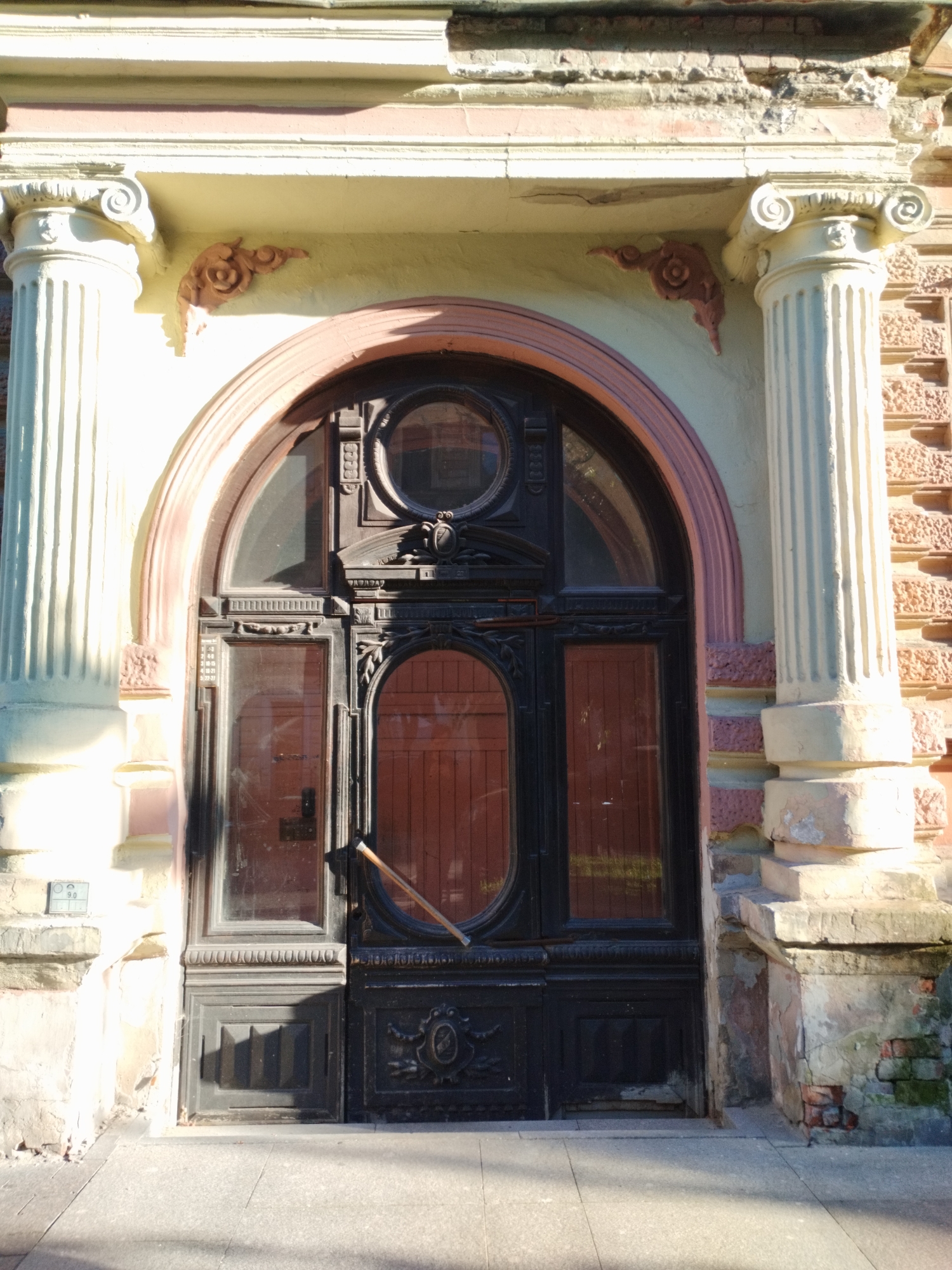 A rustic, weathered facade of a building in Saint Petersburg, Russia, is depicted, featuring a large, arched, dark wooden door framed by ornate classical columns. The door is divided into several panels, some glass and some solid wood, with a circular decorative element above the arched section. The stone columns are fluted and topped with volutes, and the building's exterior wall is constructed of brick. Decorative terracotta accents adorn the wall above the door. The scene is bathed in sunlight, casting shadows on the pavement in front of the entrance.
