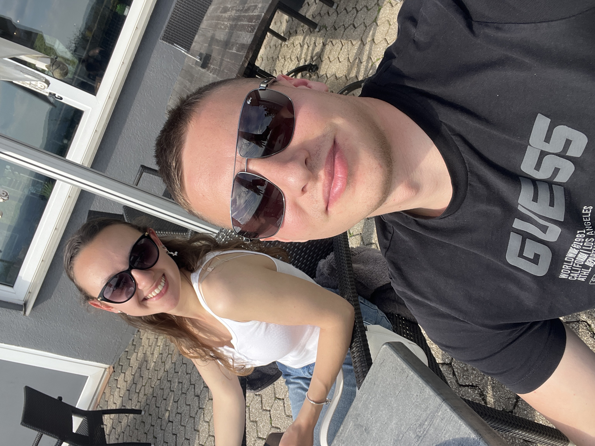 Two people, a man and a woman, are seated at an outdoor table in Saarburg, Germany. The man is wearing sunglasses and a black t-shirt with "GUESS" printed on it. The woman is wearing sunglasses and a white tank top with blue jeans. They are sitting near a building with windows and a gray exterior. The ground is paved with patterned stones.