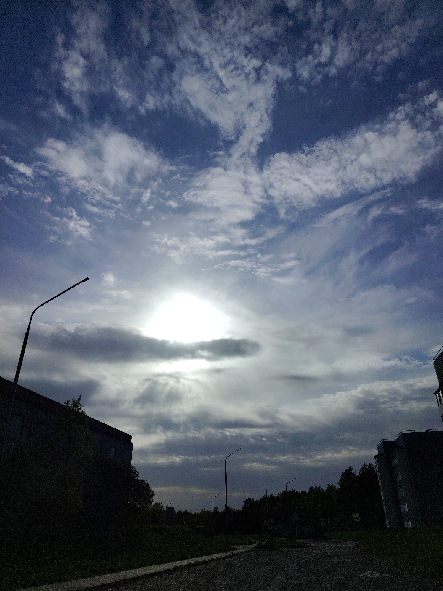 The image depicts a cloudy sky with the sun obscured behind clouds, casting rays down towards the ground. Below the sky, there's a street with streetlights and buildings on either side, silhouetted against the sky. There are trees visible in the middle ground. The location is believed to be Марковский, Russia, based on additional context.