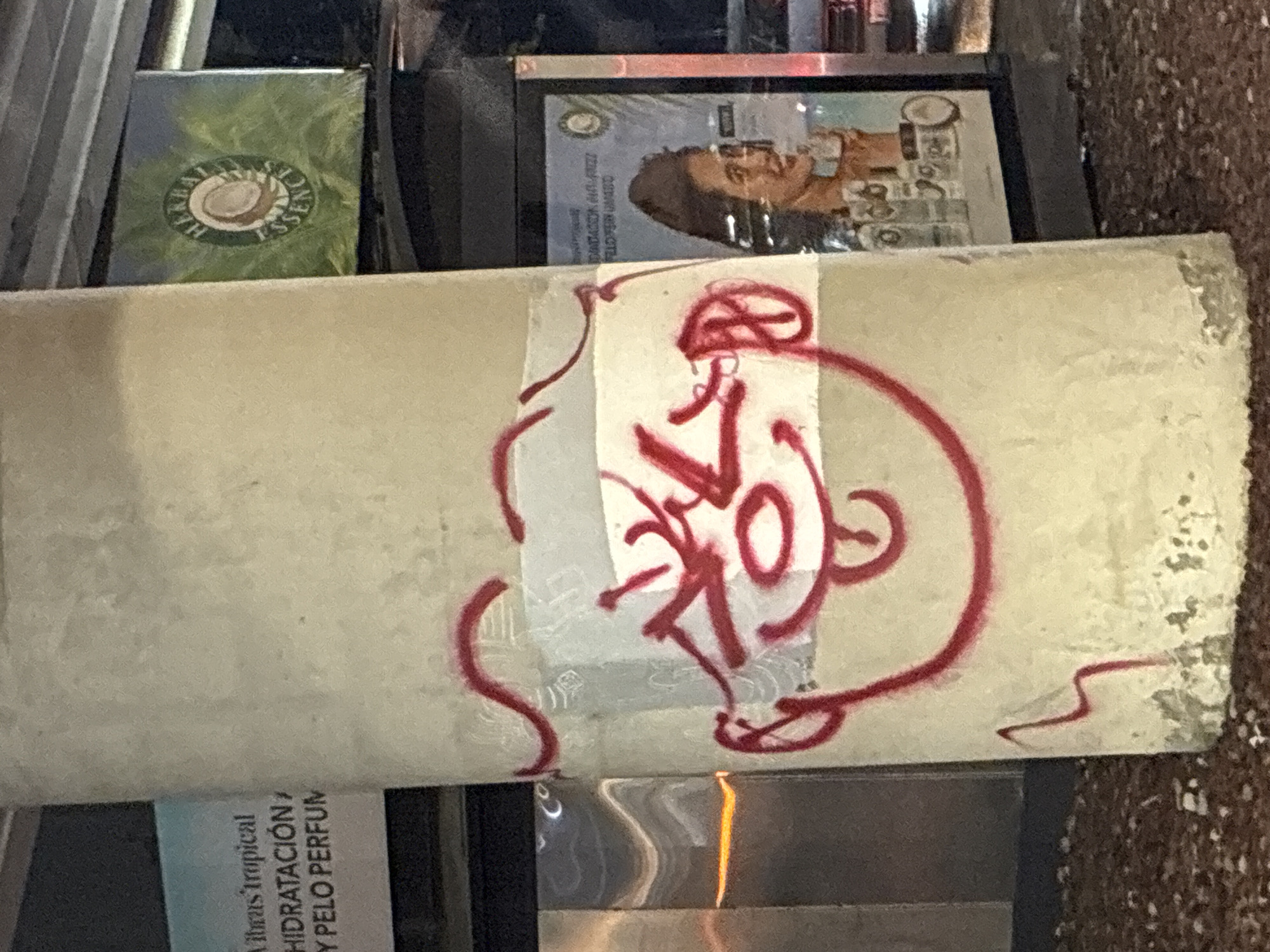 The image depicts a section of a weathered, off-white pillar or wall with red graffiti overlaid. The graffiti resembles a stylized face. Visible behind the pillar are commercial products, including a green package with a logo featuring the word "Essence," and another package with Spanish text, "Vibras tropical HIDRATACIÓN Y PELO PERFUM." Also visible is an image of a woman with text that appears to be a legal disclaimer in small font. The overall setting suggests an urban environment.