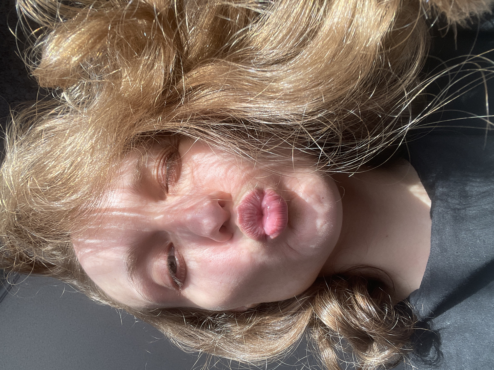 A close-up, horizontally oriented shot displays a person with fair skin and light brown to blonde wavy hair, likely taken indoors in Kuyeda, Russia. The individual, appearing to be female, is positioned with their head to the left and upper body extending to the right. Their eyes are partially closed or squinting, and their lips are puckered into a playful "kiss" or "duck face" expression. Strong, direct sunlight illuminates the face and hair, creating bright highlights on the strands and casting distinct, parallel linear shadows across the skin, possibly from window blinds or slats. The wavy hair is spread out around the head, showcasing individual strands reflecting the sunlight. The person is wearing a dark-colored, possibly black or navy, top. The background is dark and out of focus, suggesting an indoor setting. The intense sunlight indicates it is daytime with clear, sunny weather. No visible text is present. The overall impression is one of casualness and playfulness.