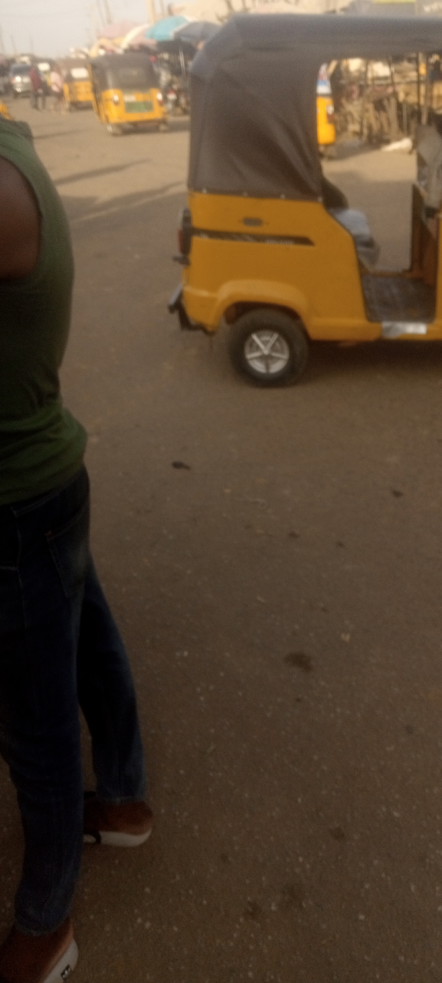 The image shows a street scene in Narayi, Nigeria. A person wearing a green sleeveless shirt and jeans stands on the left. To their right is a yellow auto-rickshaw, also known as a "keke", with a dark grey canvas roof. Other similar vehicles are visible in the background, along with indistinct buildings and people, suggesting a market or transport hub. The ground is paved and dusty.
