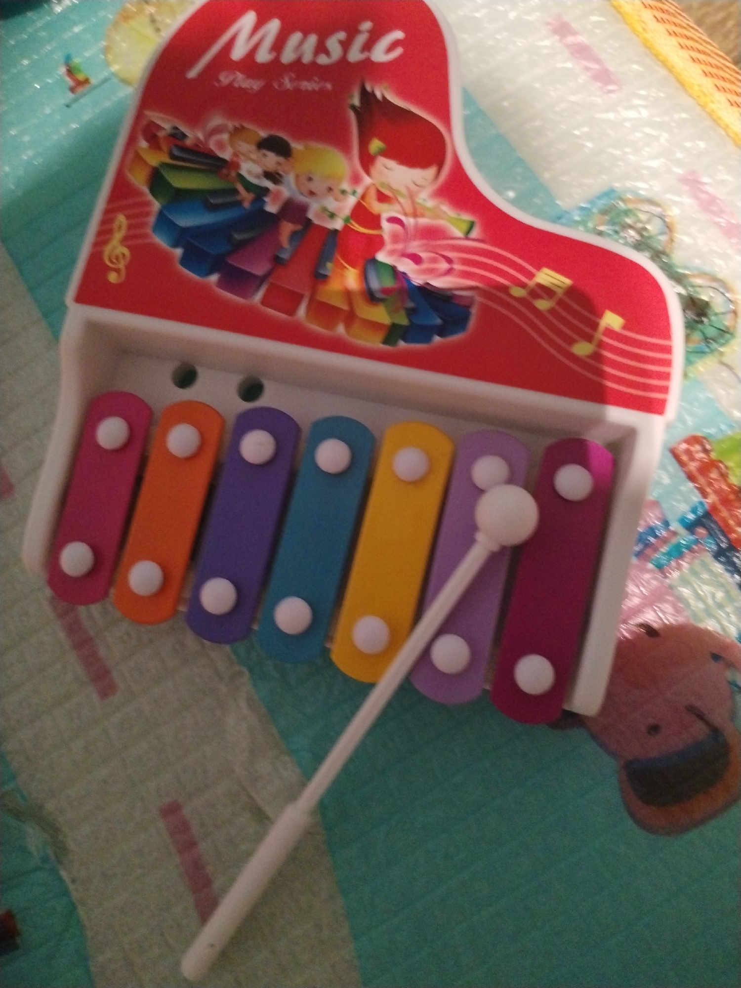 A xylophone toy with a red and white piano-shaped top is positioned in a close-up shot, likely indoors, in Nha Trang Ward, Vietnam. The instrument features a series of brightly colored metal bars: pink, orange, purple, blue, yellow, another purple, and a final pink bar. A white plastic mallet lies across the xylophone's surface. The piano top of the xylophone features an illustration of children playing musical instruments, along with musical notes. The background appears to be a patterned surface, potentially fabric, with a mix of colors including blues and greens. A child's plush toy is slightly visible to the lower right.