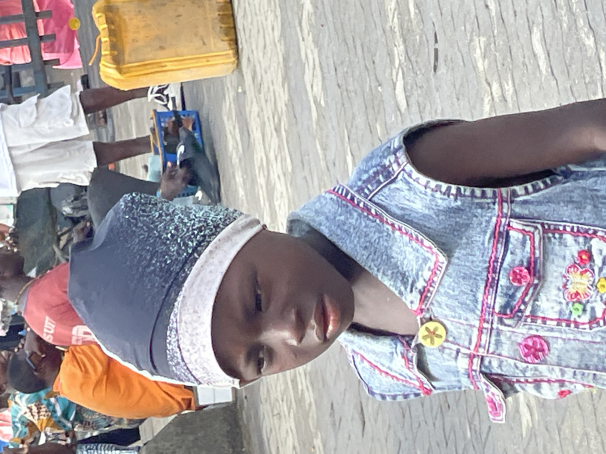 The image depicts a child with dark skin wearing a dark blue head covering with a light gray trim and a light blue denim jacket with pink trim and flower embellishments. The child is positioned in the foreground. In the background, there are several adults; one wearing a red shirt, another in orange, and a person in white shorts. There is a yellow jerrycan and blue crate also visible. The environment suggests an outdoor location, possibly a street scene in Lagos, Nigeria, inferred from the overall visual characteristics and the presence of common objects like jerrycans.