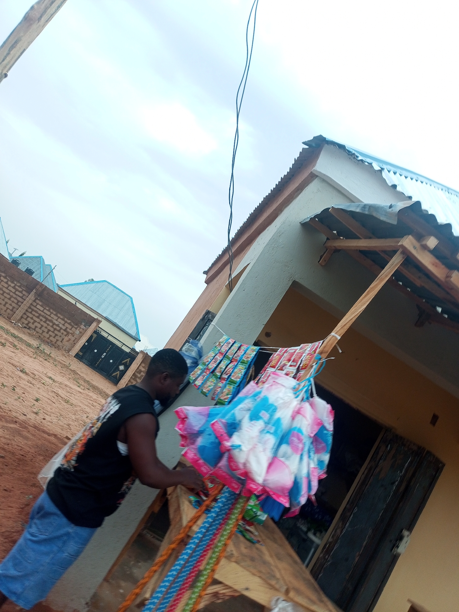 A man wearing a black t-shirt and blue shorts is standing outside of a building in Tempure, Nigeria. He is next to a display of hanging goods, possibly for sale. The building has a corrugated metal roof. In the background, there is a brick wall and another building with a blue roof. The ground is sandy.