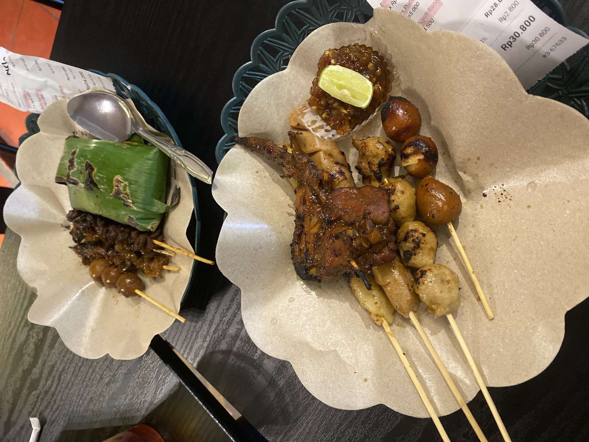 The image displays two plates of Indonesian food, likely served at a restaurant. The food consists of skewers of meat and quail eggs, with a side of rice wrapped in banana leaves. The dishes are presented on rough paper placemats with a decorative edge, placed on a dark, textured surface. In the foreground, a plate holds multiple skewers of various grilled meats, including what appears to be chicken wings and meatballs, accompanied by a small portion of sambal and a lime wedge. In the background, another plate features skewers of quail eggs and a foil-wrapped portion of rice. A spoon rests on the edge of the background plate. A receipt with Indonesian currency denominations (Rp) is partially visible at the bottom right, suggesting a transaction. The overall setting appears to be a casual dining environment.