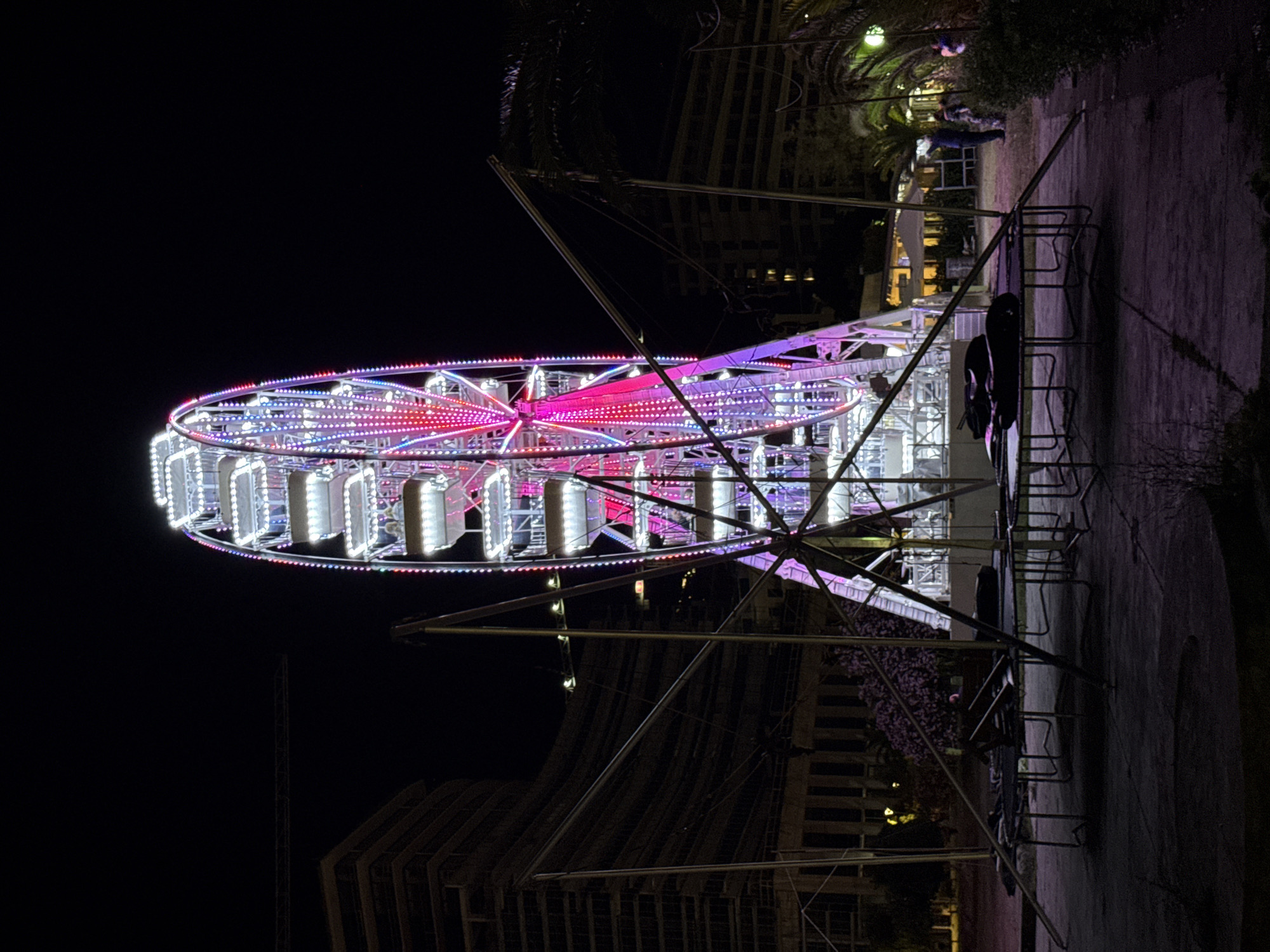 An illuminated Ferris wheel stands tall at night in Budva, Montenegro, likely in a park or amusement area. The wheel is adorned with a vibrant display of pink, purple, and white lights, outlining its structure and cabins. In the foreground, several trampolines are set up, some with dark objects, possibly cushions or mats, lying on them. The ground appears to be a paved area or dark soil.
In the background, multi-story buildings with illuminated windows are visible, suggesting an urban setting. To the right, palm trees and other foliage frame the scene, with some areas of the background showing distant lights from buildings or possibly a promenade. A few people are visible in the middle ground, near a seated area with umbrellas. One person appears to be pointing towards something out of frame, while another stands next to them. The overall atmosphere is that of a nighttime recreational space.