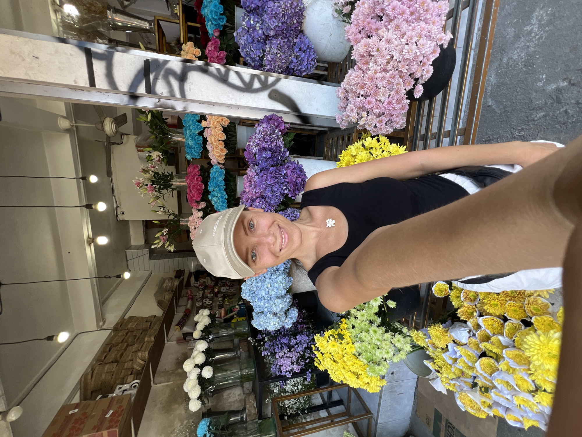 A woman is taking a selfie in a flower shop. She is wearing a black tank top, a light-colored baseball cap, and a white necklace with a small floral pendant. She is smiling at the camera. The flower shop is filled with an array of colorful flowers, including hydrangeas, lilies, and chrysanthemums. The flowers are arranged in buckets and vases. The shop has a metal roof, and there are shelves and boxes visible in the background. Based on the metadata, the location is Peliatan, Indonesia.