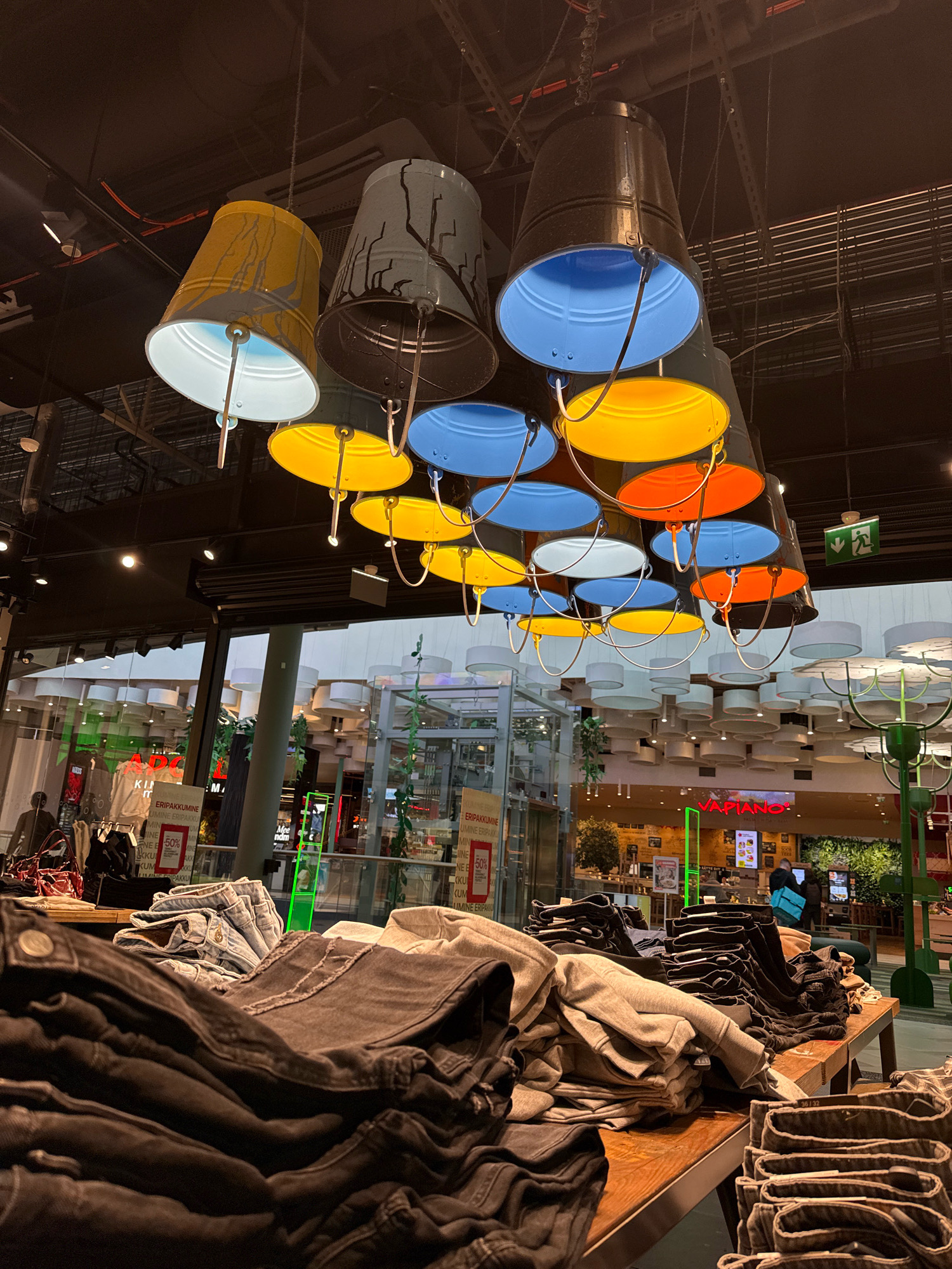 This image depicts the interior of a retail store, likely a clothing store given the display of folded garments in the foreground. The store is illuminated by a unique lighting installation featuring multiple buckets of various colors (yellow, blue, orange, white, and dark gray) suspended from the ceiling, serving as lampshades. In the background, through a glass partition, a dining establishment with visible signage ("Vapiano") and people can be seen. The presence of the Vapiano restaurant suggests the location is likely a shopping mall or a large retail complex. Considering the context of Tallinn, Estonia, this scene could be situated within a mall such as Ülemiste Keskus or Rocca al Mare Keskus, which often house such dining and retail establishments. There are several racks of folded clothing, primarily jeans, on display tables in the foreground and midground. People are present in the background, some appearing to be customers or employees within the dining area. The environment appears to be a modern indoor commercial space.
