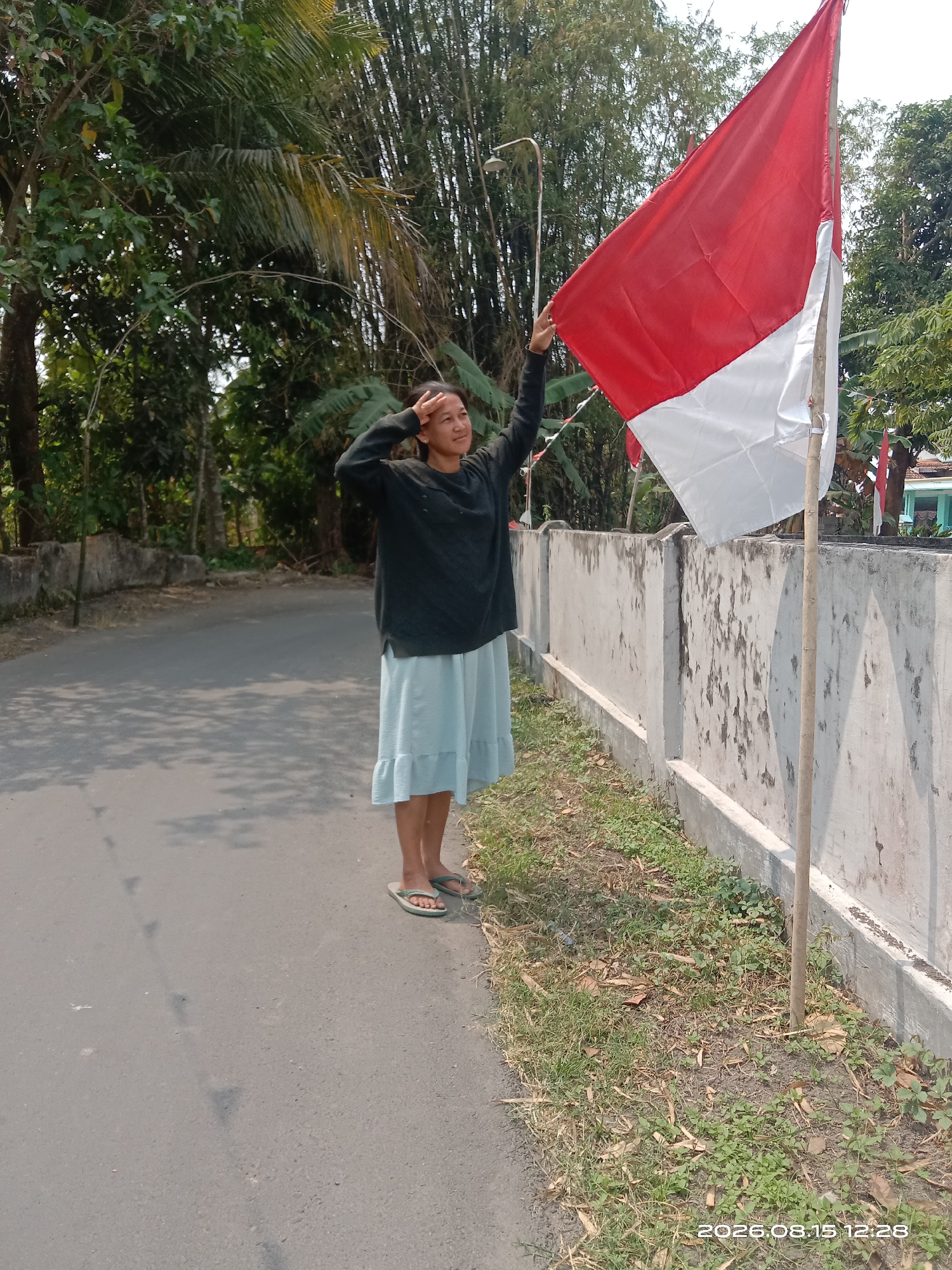 A young woman stands on the side of a paved road, performing a salute. She wears a dark long-sleeved shirt and a light blue ruffled skirt, with flip-flops on her feet. In her right hand, she holds a red and white Indonesian flag attached to a wooden pole. The flag is partially unfurled, with the red section at the top and the white section below. The woman's head is turned to her right, and her gaze is directed upwards.
Behind her, a white concrete wall runs along the edge of the road, bordered by grass and dry leaves. Further beyond the wall, a dense stand of bamboo trees rises high into the sky, interspersed with other foliage. The road is paved and appears to be a rural or suburban street, with shadows cast on its surface from unseen trees. In the far background, glimpses of buildings and more trees can be seen. The overall scene suggests a patriotic display or commemoration, likely taking place outdoors in Indonesia. The timestamp in the lower right corner indicates the image was captured on August 15, 2026, at 12:28.