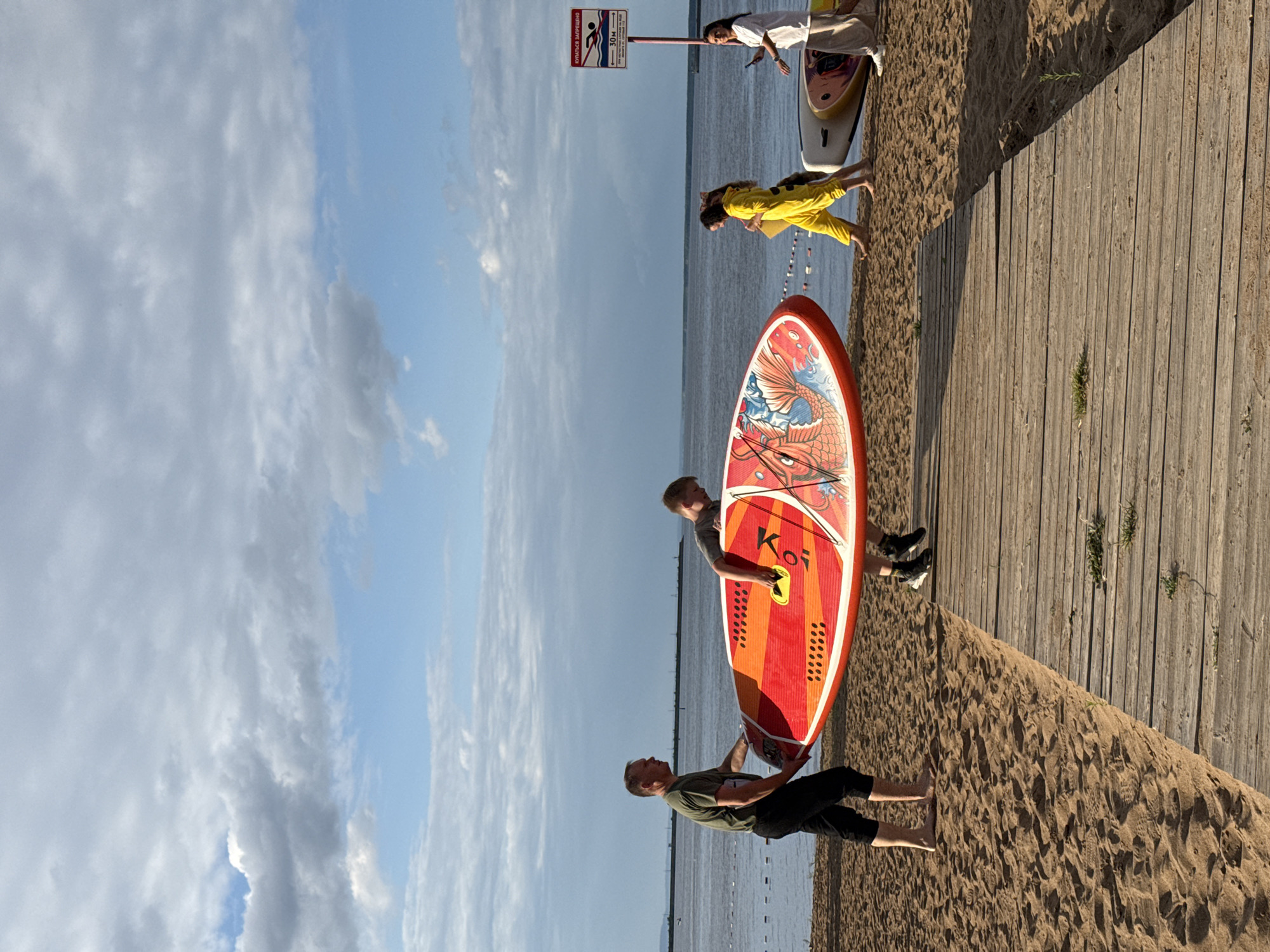 A group of people are on a sandy beach near a body of water, possibly the Kama River, in Chaikovsky, Russia. In the foreground, two men are carrying a large, colorful paddleboard. To the right, two women are walking and standing near other paddleboards. One woman is wearing a bright yellow outfit, and the other is in white. A red and white sign is posted on a pole behind them. The sky is partly cloudy, and the overall lighting suggests it is daytime. A wooden walkway leads across the sand.
