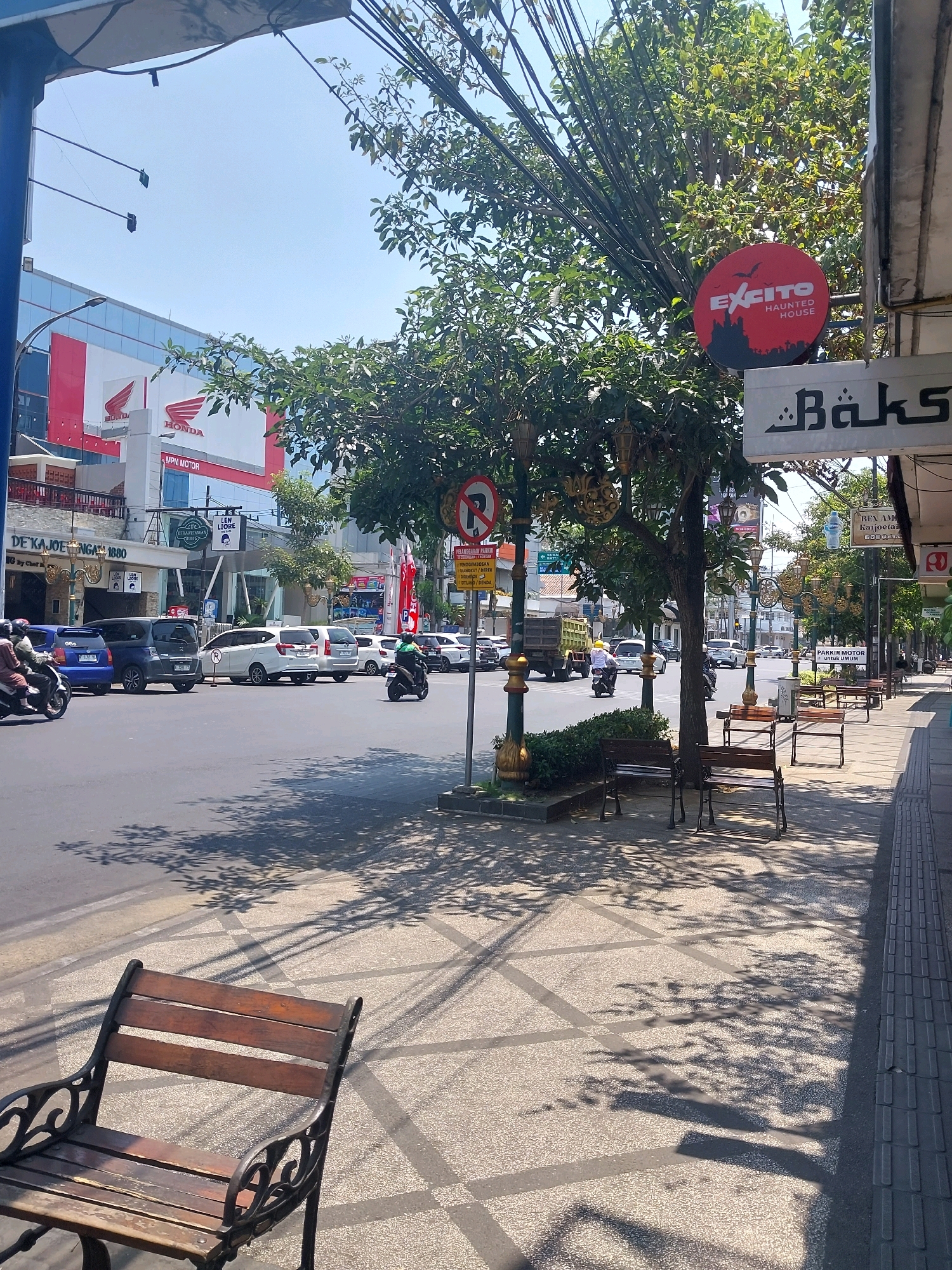 This image depicts a street scene in Malang, Indonesia, likely during daylight hours given the strong shadows. On the left side of the road, a building with a red and white facade, featuring the "HONDA" logo, is visible. Across the street, a sidewalk area is lined with benches and trees, casting dappled shadows on the patterned pavement. Several vehicles, including cars and motorcycles, are present on the road, suggesting moderate traffic. A sign with "Bäks" written on it is visible above a storefront. Another circular red sign with a silhouette of a castle and bats reads "EXCITE HAUNTED HOUSE". The overall environment appears to be an urban commercial area.