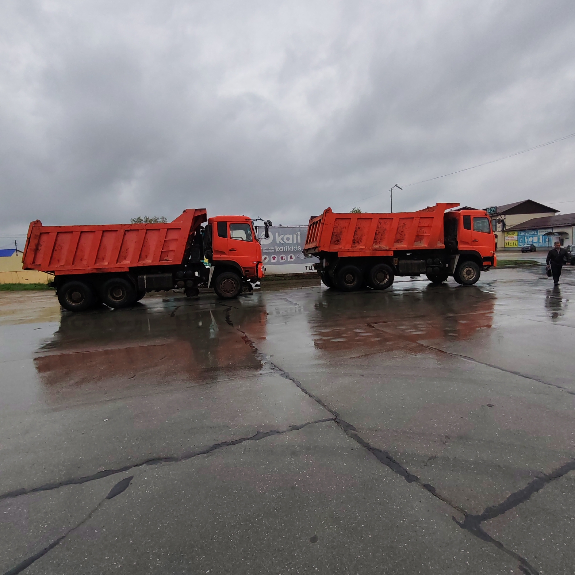 The image shows two orange dump trucks parked on a wet asphalt surface with cracks and puddles. The sky is overcast. A man is walking to the right of the frame, and in the background, there are buildings and signage in Russian, including a sign for "kari kids." The location is likely Chaikovsky, Russia, based on the signage.