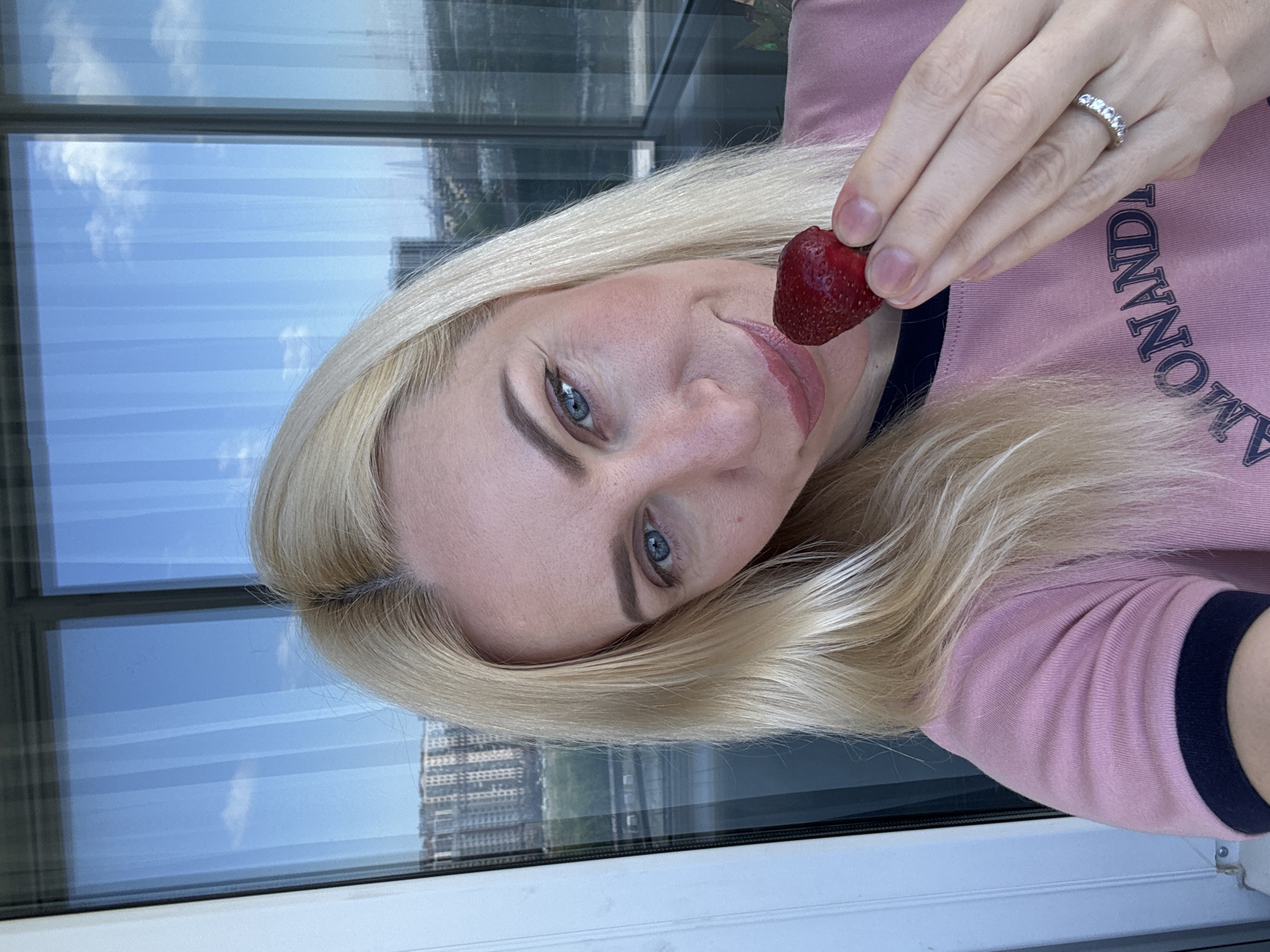 A woman with blonde hair and blue eyes is holding a strawberry up to her mouth, looking at the camera. She is wearing a pink t-shirt with dark trim around the collar. The background appears to be a city skyline seen through a large window, possibly from an apartment building. The sky is blue with some white clouds, and there are reflections in the glass. The woman is wearing a ring with several small stones on her left ring finger. The overall scene is brightly lit, suggesting it is daytime.